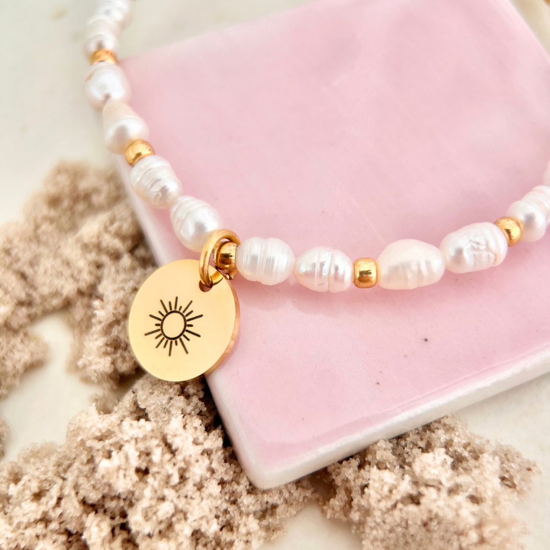 Nahaufnahme eines Perlenarmbands mit unregelmäßigen weißen Süßwasserperlen, kleinen goldenen Elementen und rundem Anhänger mit eingravierter Sonne auf einem rosafarbenen Stein