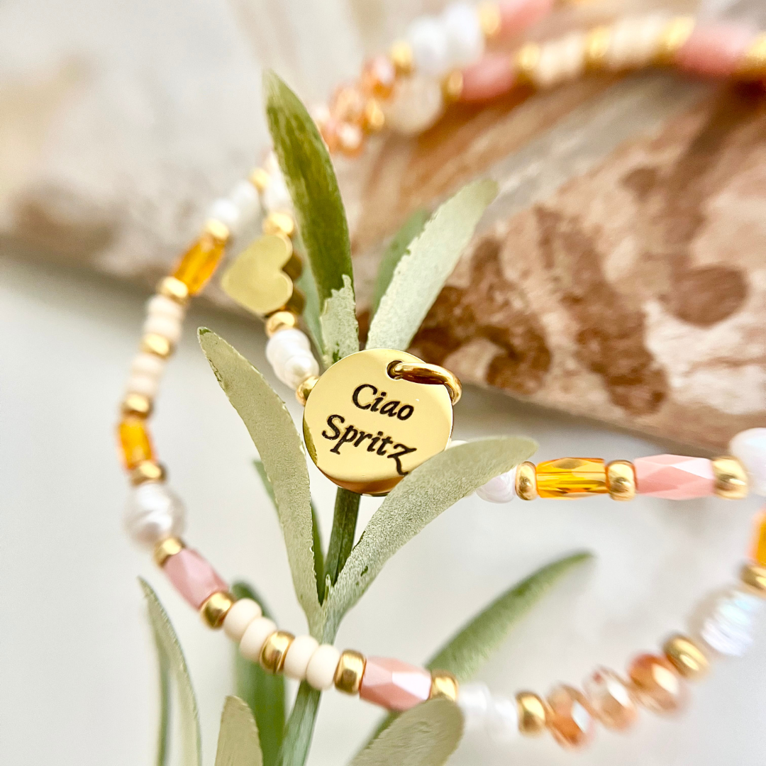 Sommerliches Perlenarmband in Orange- und Weißtönen mit goldenem „Ciao Spritz“-Gravuranhänger, fotografiert im Detail. Holy Aperoli Armband.