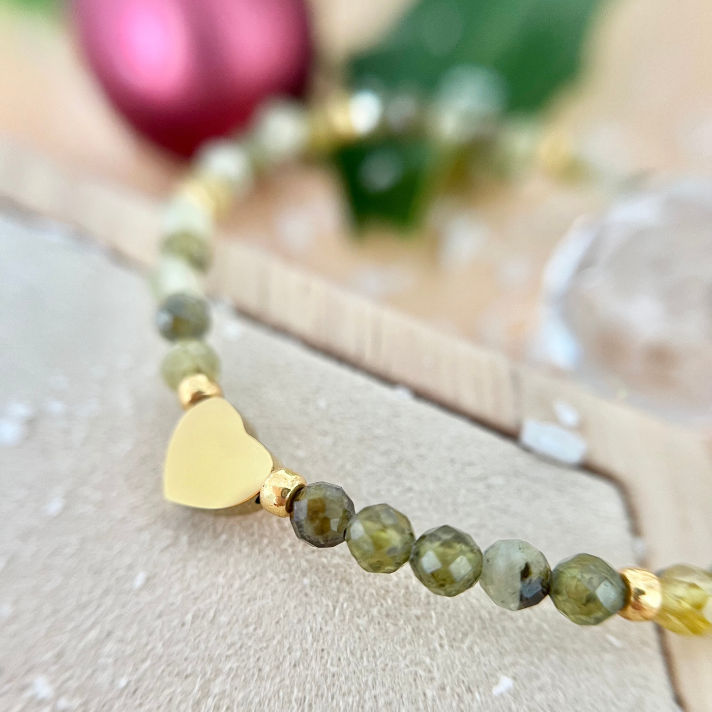 Detailansicht des Armbands mit grün schimmernden Natursteinperlen und einem goldfarbenen Herz-Anhänger aus Edelstahl, unscharfer Hintergrund mit Weihnachtsdeko.