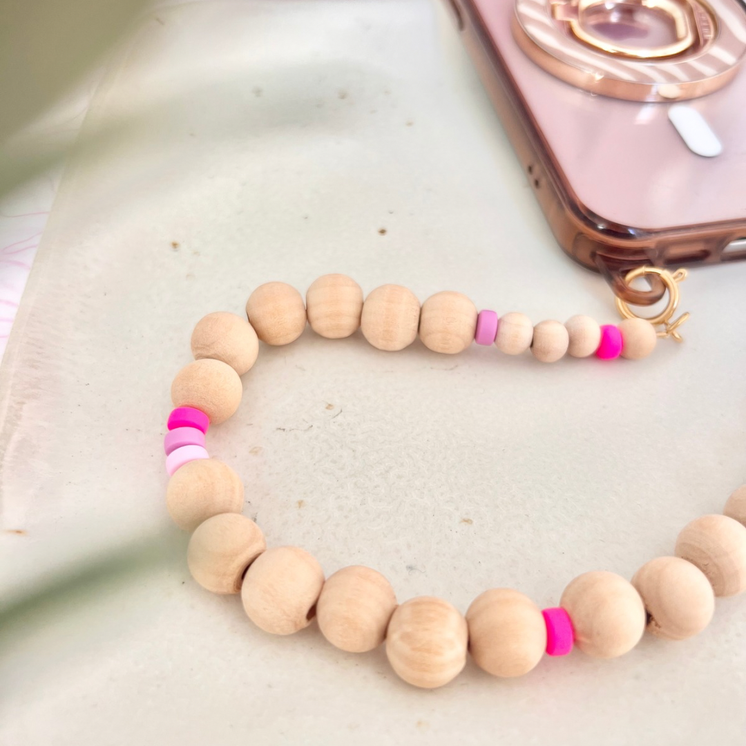 Draufsicht auf eine im Workshop gefertigte handgemachte Perlen-Handykette aus runden Holzperlen in Naturbeige mit kleinen Silikonperlen in Pink- und Lilatönen, befestigt an einem Smartphone mit goldfarbenem Karabinerverschluss auf hellem Untergrund.
