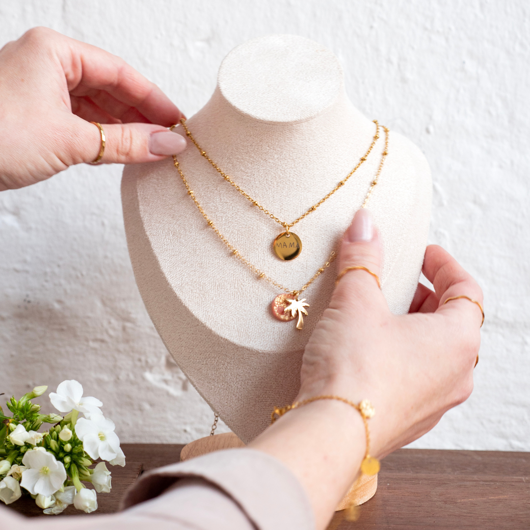 Nahaufnahme einer Schmuckbüste mit zwei goldenen Ketten, eine mit rundem Gravur-Anhänger „MAMA“, die andere mit Palmenmotiv, gehalten von Händen mit goldenen Ringen und Armband, daneben weiße Blüten.