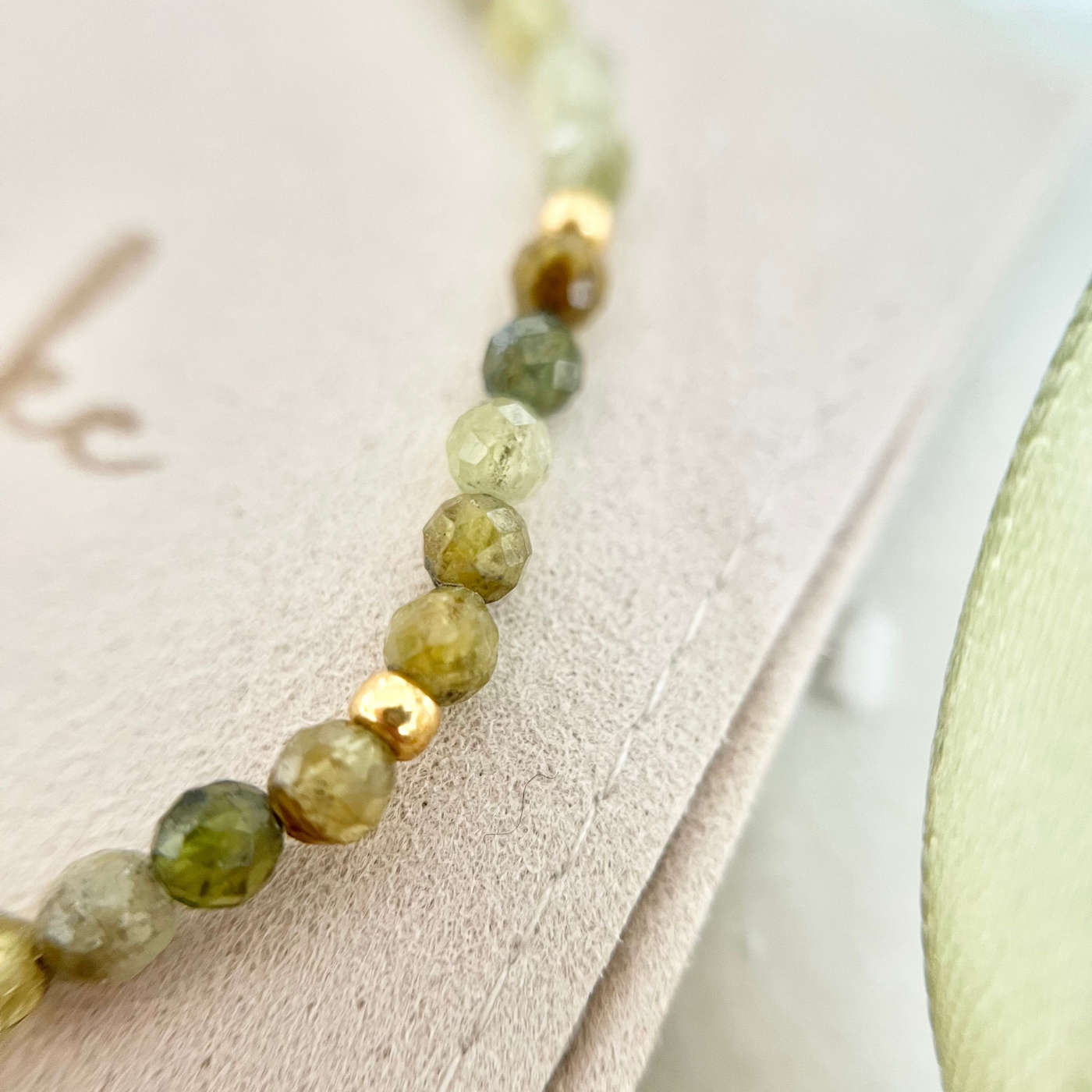 Nahaufnahme eines Armbands mit facettierten Natursteinperlen in verschiedenen Grüntönen, kombiniert mit goldfarbenen Zwischenelementen. Das Schmuckstück liegt auf einem beigen Beutel mit „Danke“-Aufschrift – stilvolles Geschenk für Weihnachten oder besondere Anlässe.