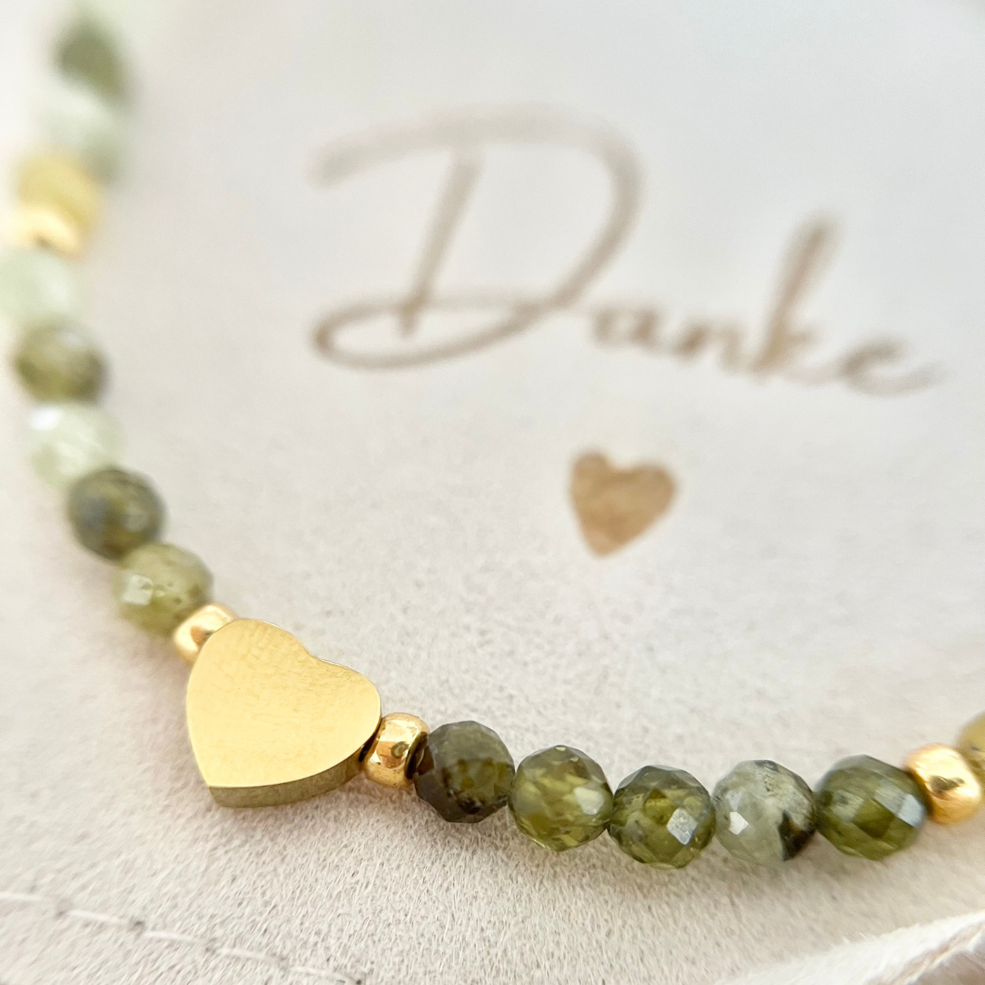 Nahaufnahme eines handgefertigten Perlenarmbands aus grünen Natursteinperlen mit vergoldetem Herz-Anhänger. Das Armband liegt auf einem Schmuckbeutel mit der Aufschrift „Danke“.