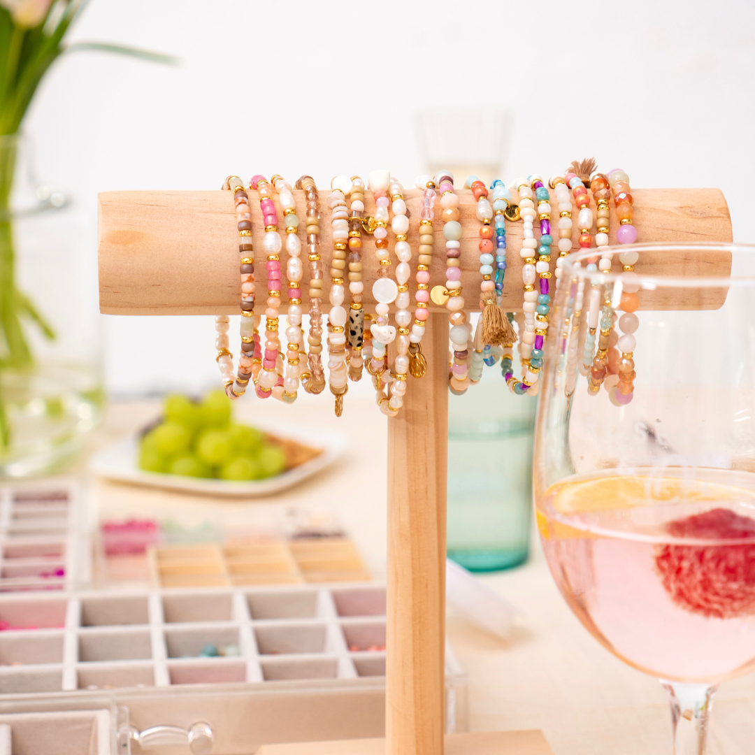 Bunte Perlenarmbänder auf Holzständer als Inspiration aus dem Perlenschmuck Workshop in Wuppertal – Armband Workshop für Anfänger als kreative Me-Time mit kleinen bunten Perlen, Süßwasserperlen und goldenen Anhängern.