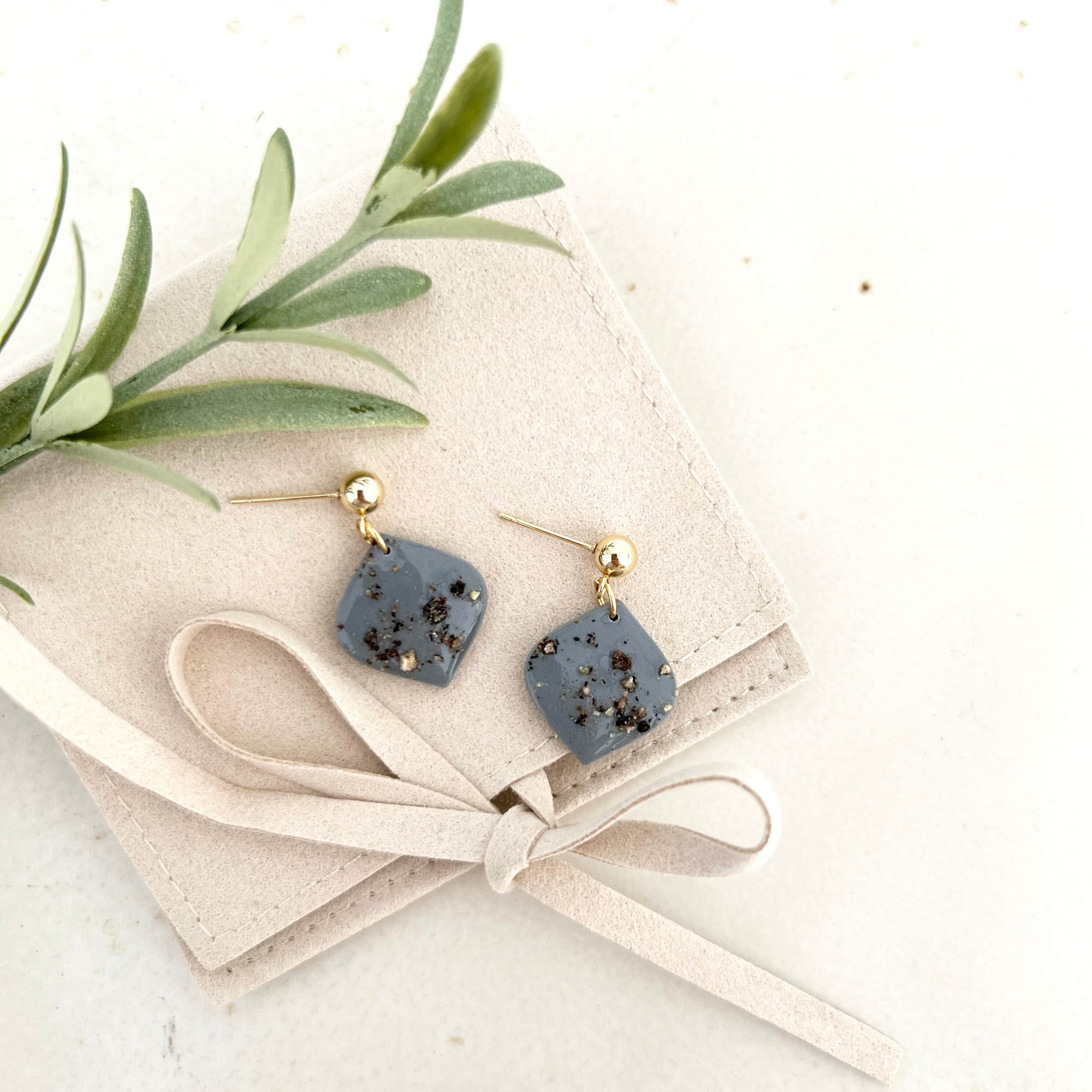 Zwei Paar Ohrringe PIMIENTA auf einer Tasche drapiert, die goldfarbenen und silberfarbenen Ohrstecker aus Polymer Clay und Edelstahl