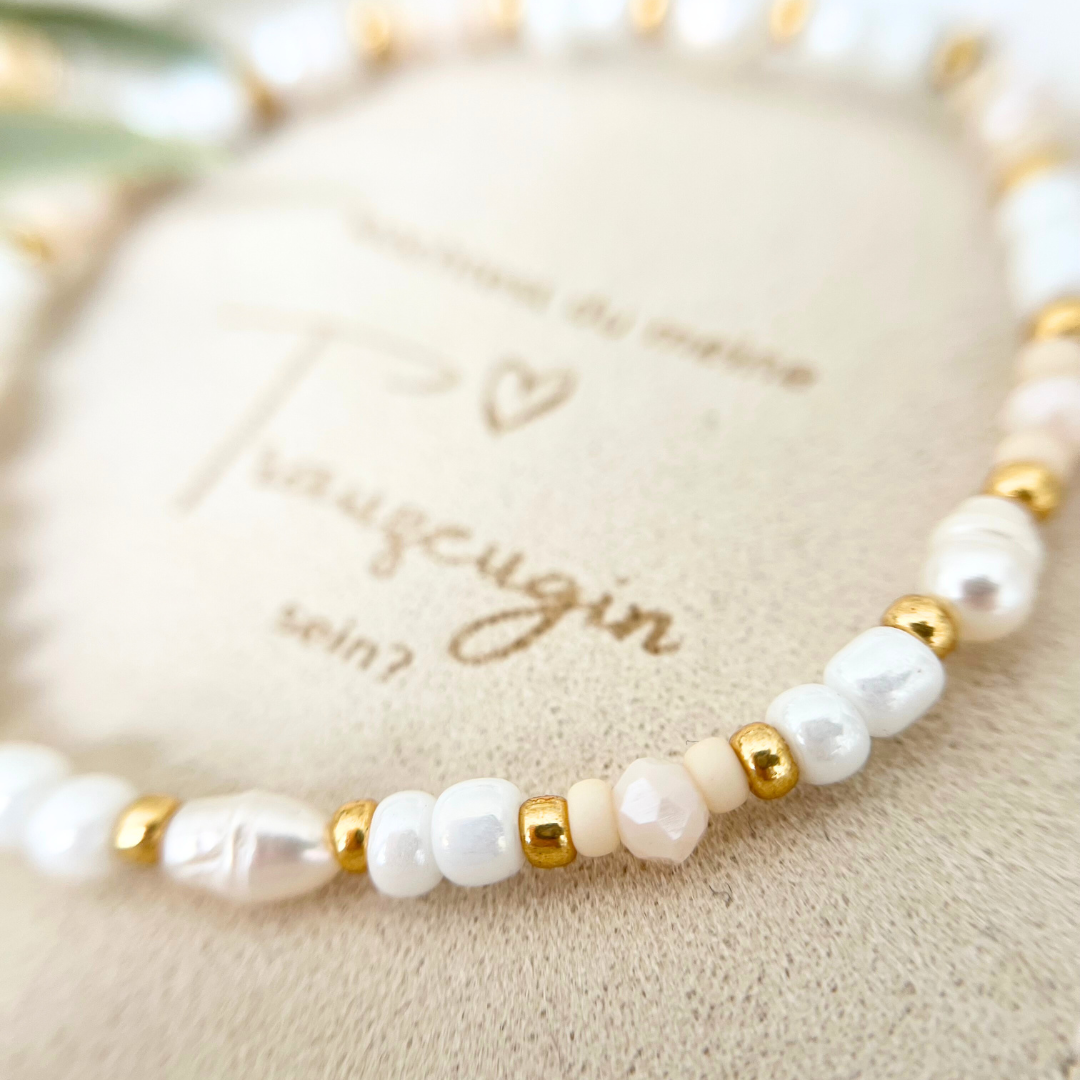 Detailansicht von Trauzeugin Schmuck – filigranes Armband mit glänzenden Perlen in Weiß, Nude und Gold, personalisierter Gravuranhänger mit Herzsymbol, auf beigem Schmuckbeutel mit gestickter Aufschrift „Trauzeugin“