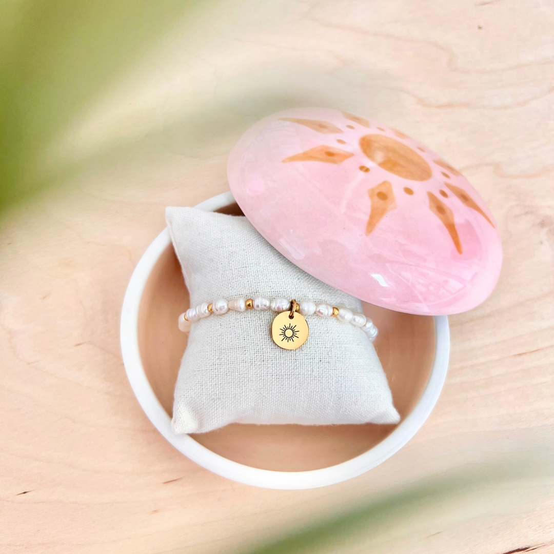 Perlenarmband mit kleinen Süßwasserperlen und goldenem Sonnenanhänger auf Schmuckkissen in runder Dose