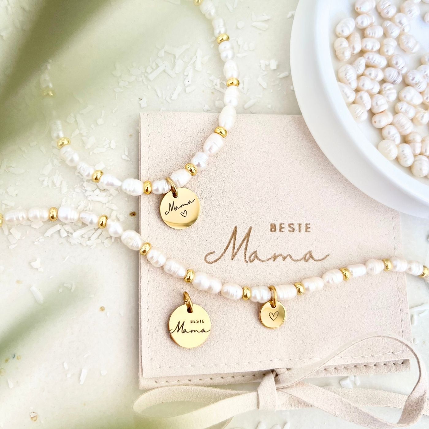 Armband aus weißen Süßwasserperlen mit goldenen Elementen und Herz-Anhänger, präsentiert auf Schmucktäschchen „Beste Mama“.