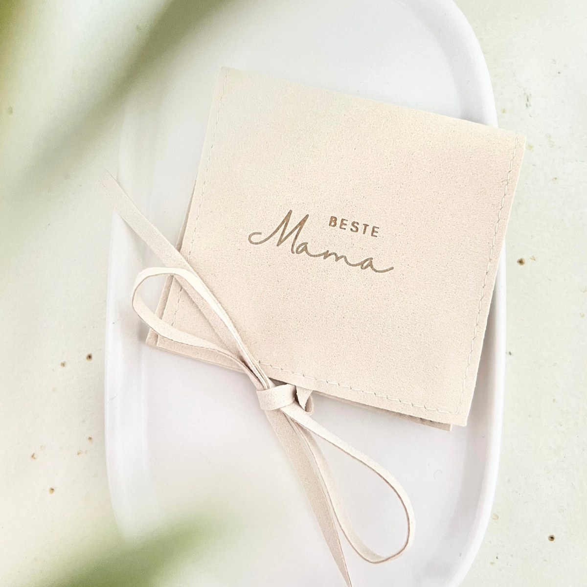 Beigefarbener Schmuckbeutel mit Schleifenband und goldener Aufschrift „Beste Mama“, liegend auf einem weißen Teller mit grünen Blättern im Vordergrund – Geschenkverpackung für Schmuck.