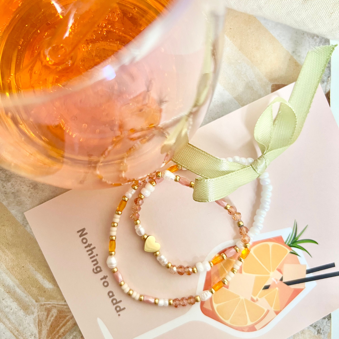 Perlenarmbänder in Orange und Weiß neben einem Weinglas gefüllt mit Aperol, stimmungsvolle Sommeraufnahme. Holy Aperoli Armband.