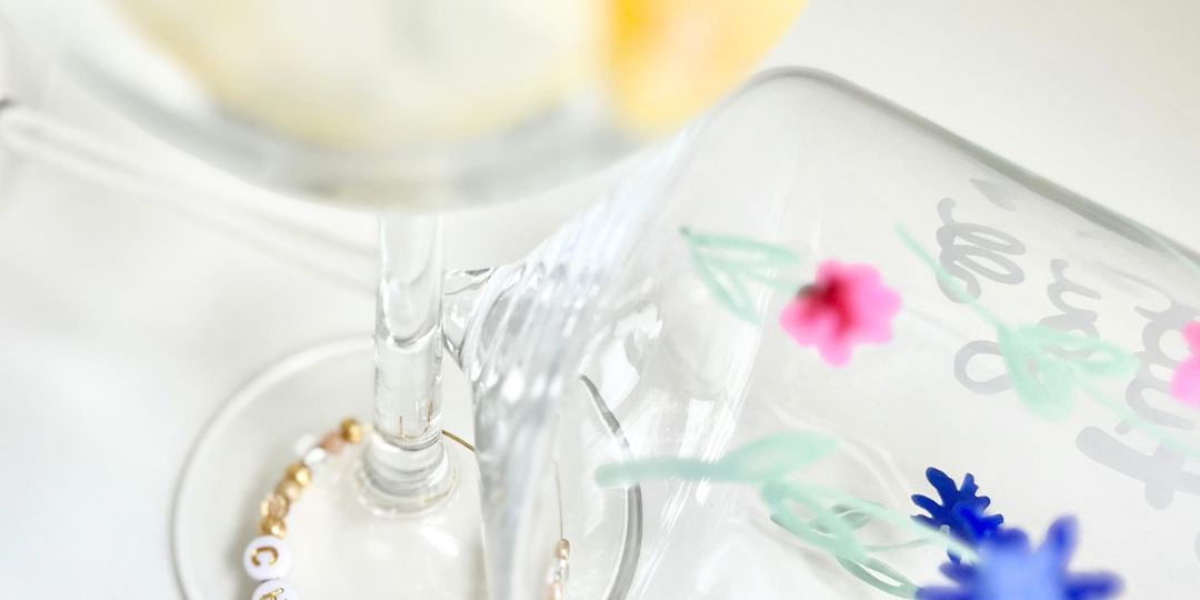 Zwei kreativ bemalte Weingläser: eines mit filigraner und floraler Malerei in pink, dunkelblau und pastelligem Salbeigrün, das andere mit gemalter Zitrone – ergänzt durch einen personalisierten Glasmarkierer mit ‚CHEERS‘ Buchstabenperlen und rosa Blüten Charm; DIY Projekt aus dem Glass Painting Workshop in Wuppertal und Wülfrath Düssel.