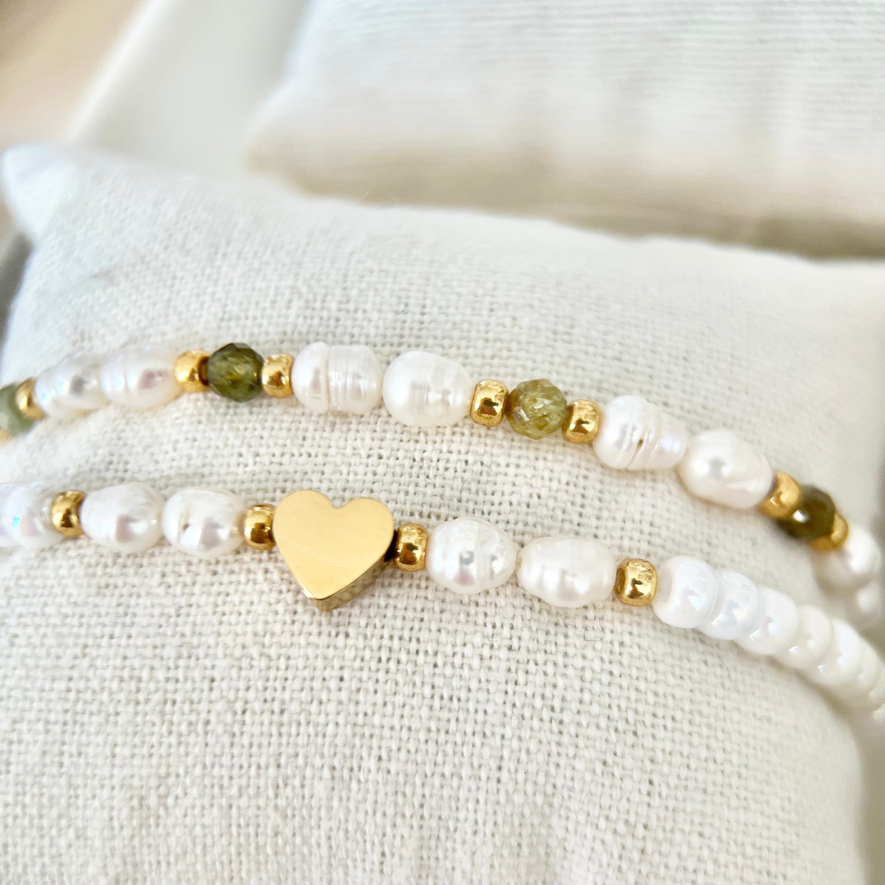 Nahaufnahme von zwei Armbändern mit weißen Süßwasserperlen, grünen Natursteinen und goldenen Herz-Details auf beigem Stoffkissen.