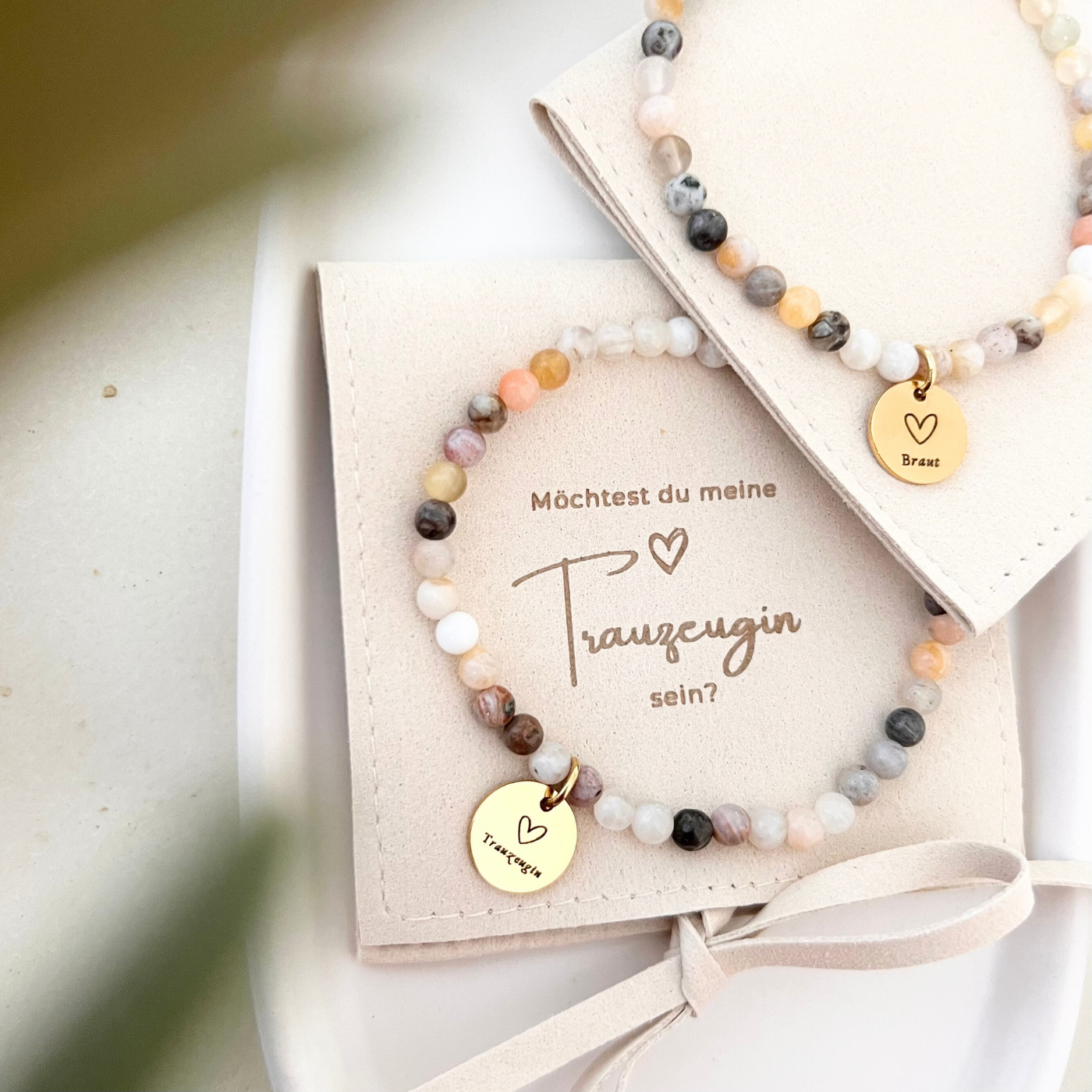 Detailaufnahme eines Perlenarmbands mit weißen und beigen Natursteinen, goldfarbenem Anhänger mit Herzsymbol und Schriftzug „Braut“, dekoriert mit grünem Blatt.