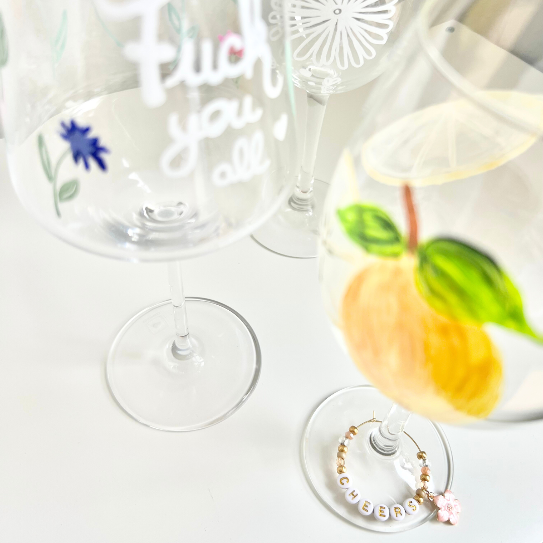 Drei bemalte Weingläser mit individuellen Designs: handgeschriebener Lettering Schriftzug, weißes Boho Blütenmuster und gemalte Zitrone mit Blättern – dazu ein personalisierter Weinglas Charm mit ‚CHEERS‘-Buchstabenperlen und Blüten-Anhänger; kreative Ergebnisse aus dem Glass Painting Workshop in Wuppertal