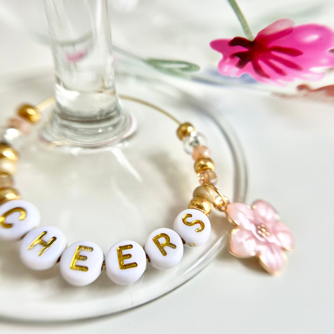 Detailaufnahme eines bemalten Weinglases mit pinker Blume und personalisiertem Metallring mit Perlen – Buchstabenperlen in Weiß mit goldener Aufschrift ‚CHEERS‘, kombiniert mit rosa Glasperlen, goldenen Akzenten und einerm Blüten Anhänger; DIY Projekt aus dem Glass Painting Workshop in Wülfrath