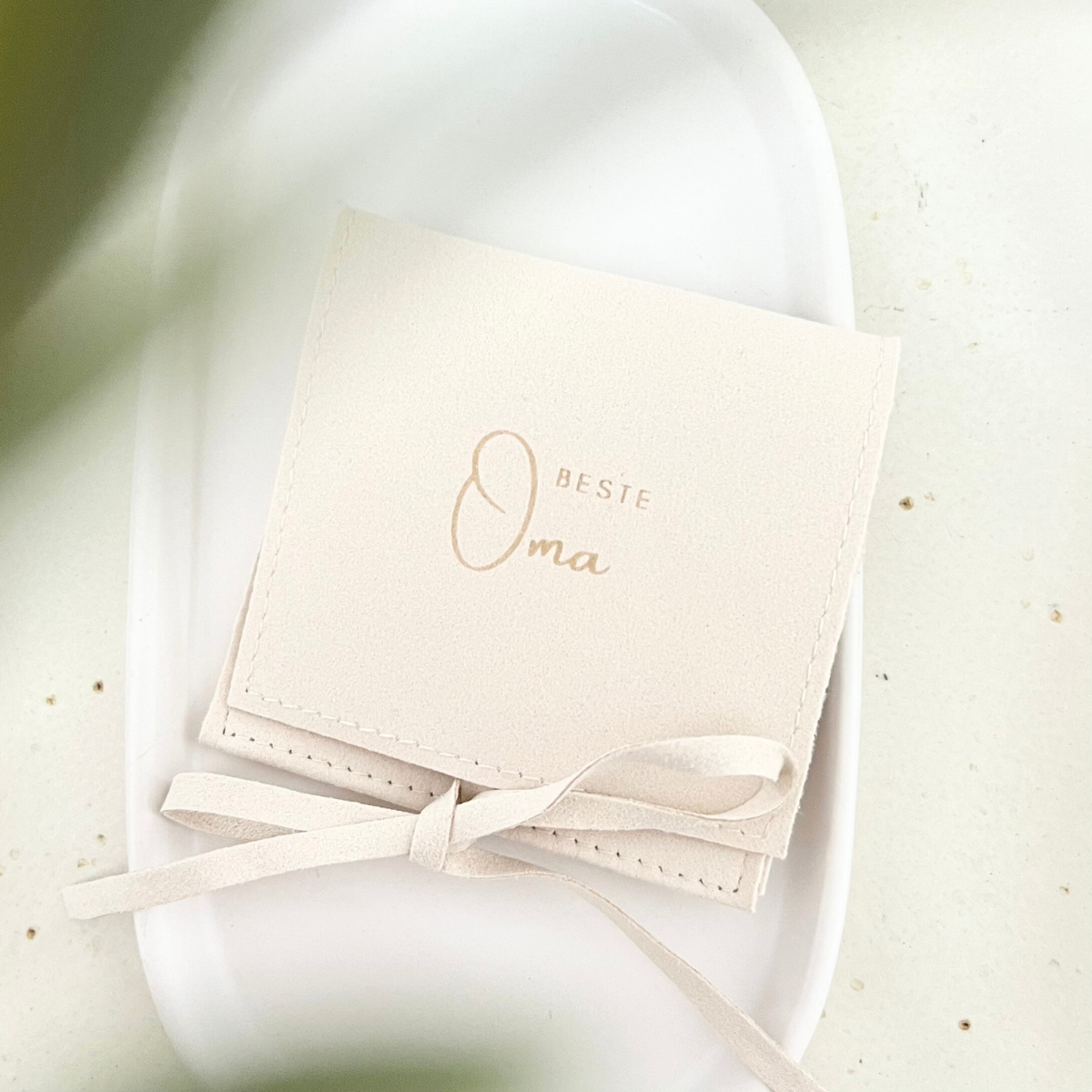 Nahaufnahme eines beigen Schmuckbeutels aus Mikrofaser mit Schleife und der Aufschrift „Beste Oma“, liegend auf heller Keramikschale – Geschenkverpackung für Schmuck