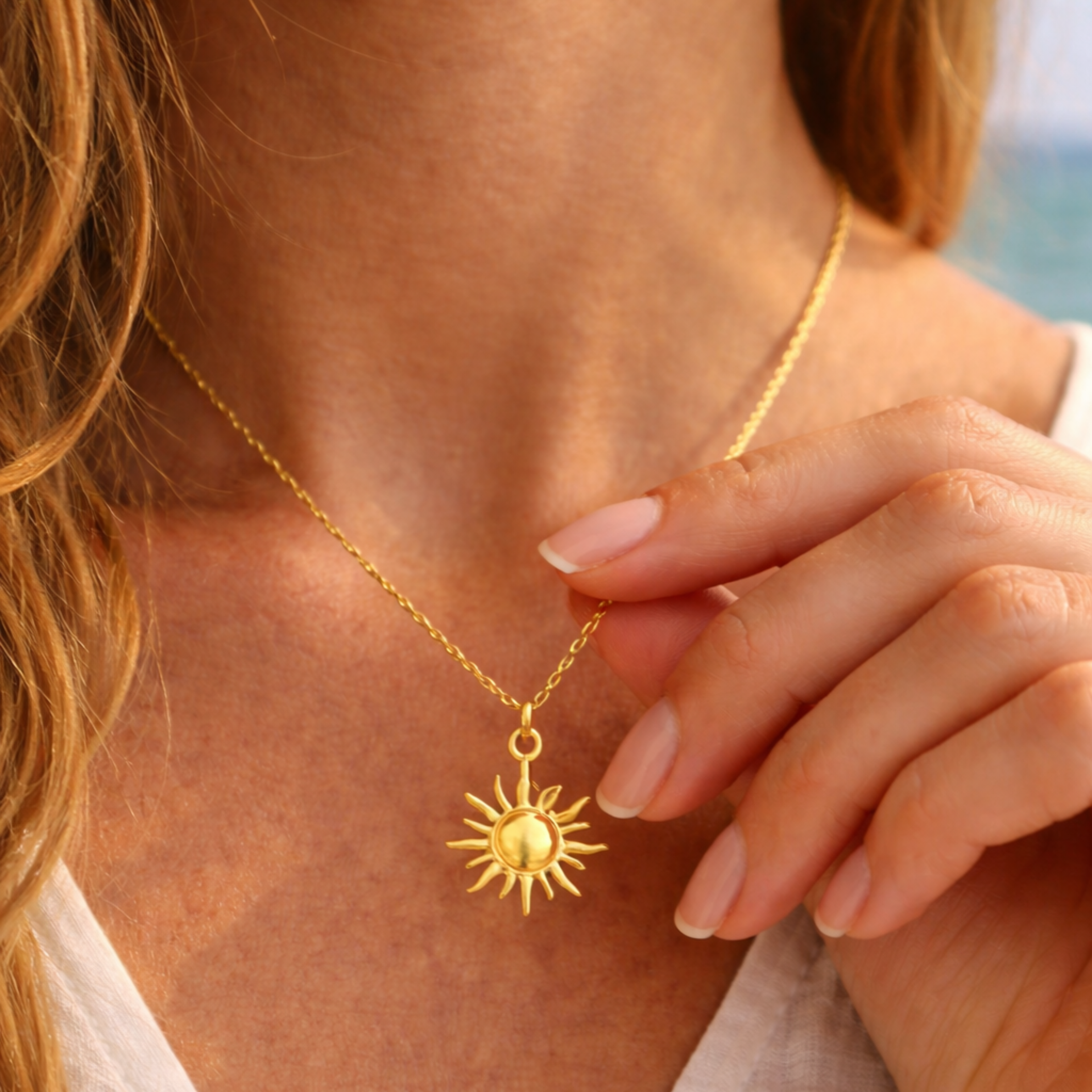 Goldene Sonnenkette aus Edelstahl wird von einer Frauenhand am Hals gehalten, filigrane Kette mit glänzendem Sonnenanhänger auf leicht gebräunter Haut, getragen zu weißem Oberteil