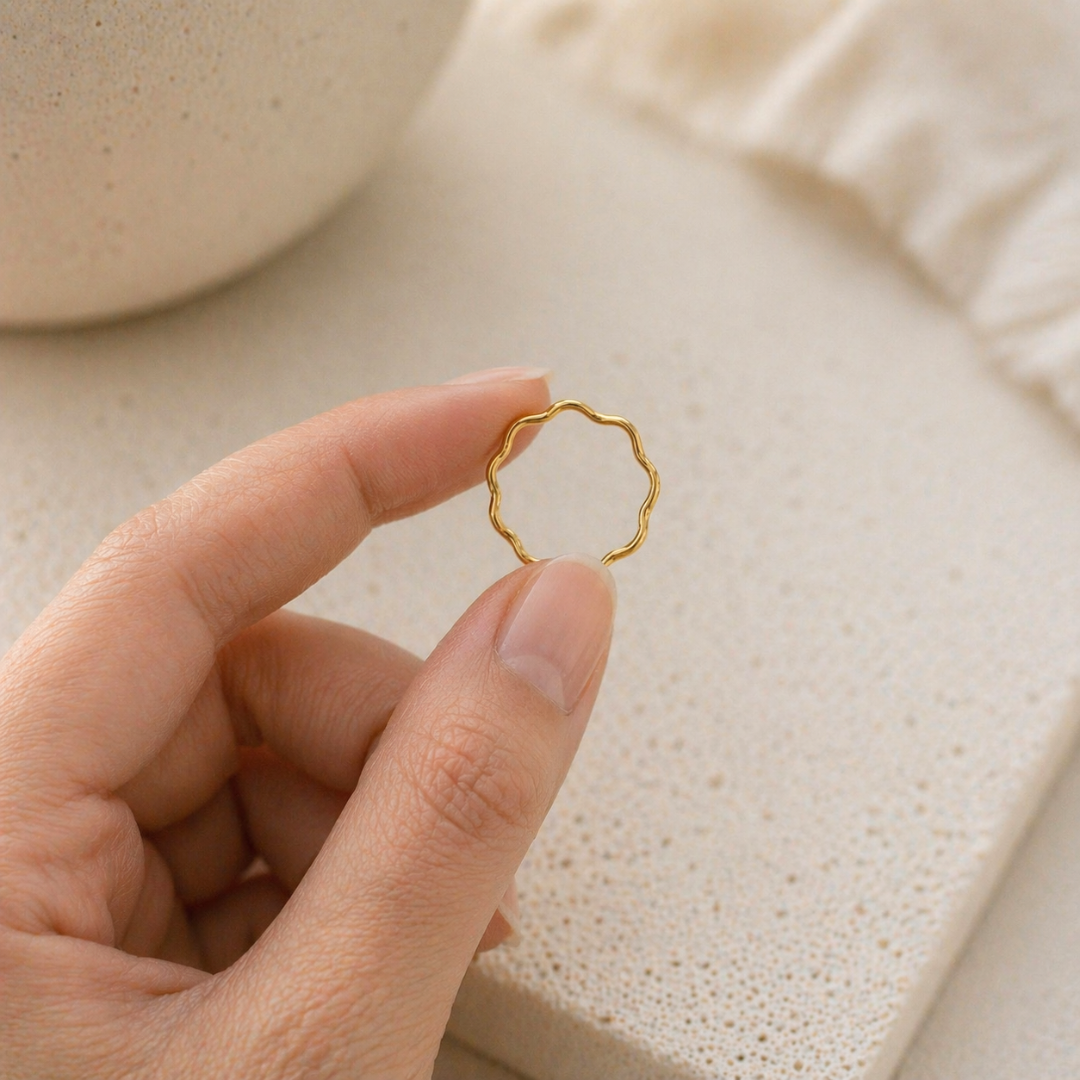 Zarter goldener Wellenring aus Edelstahl wird zwischen zwei Fingern gehalten, natürliche Hautstruktur, heller Hintergrund, minimalistisches Schmuckbild