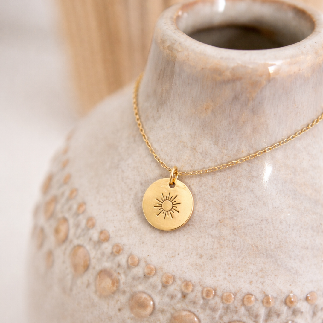 Feine goldene Halskette aus Edelstahl mit kleinem runden Anhänger und eingravierter Sonne, dekorativ auf Vase drapiert, ideal als sommerliches Schmuckstück oder Geschenk