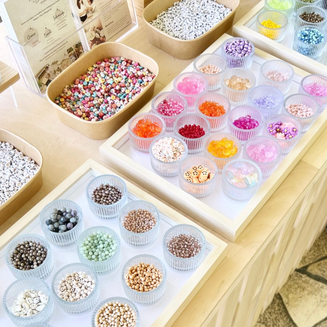 Bunte Perlen in verschiedenen Farben und Materialien, sortiert in kleinen Glasschälchen auf Tabletts. Im Hintergrund weitere Schalen mit Buchstabenperlen und farbigen Rocailles. Die Auswahl ist Teil des DIY Perlen Workshops bei Rosemary & Olive in Wuppertal