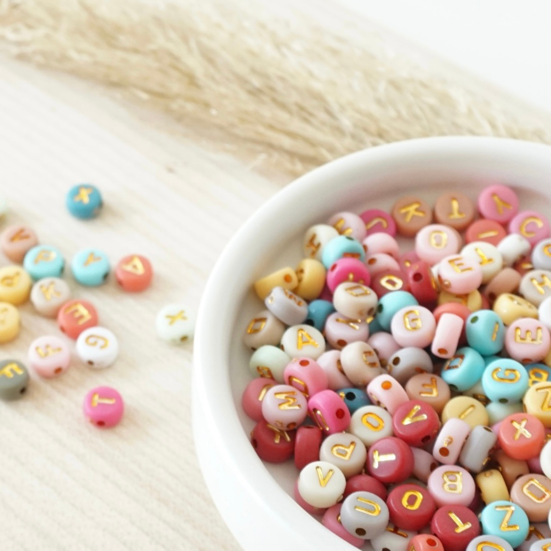 Bunte Buchstabenperlen in Pastellfarben mit goldener Schrift in einer weißen Schale – ideal zum Personalisieren von Armbändern im DIY Schmuck Workshop bei Rosemary & Olive in Wuppertal. Perfekt für Namen, Botschaften oder liebevolle Geschenkideen.
