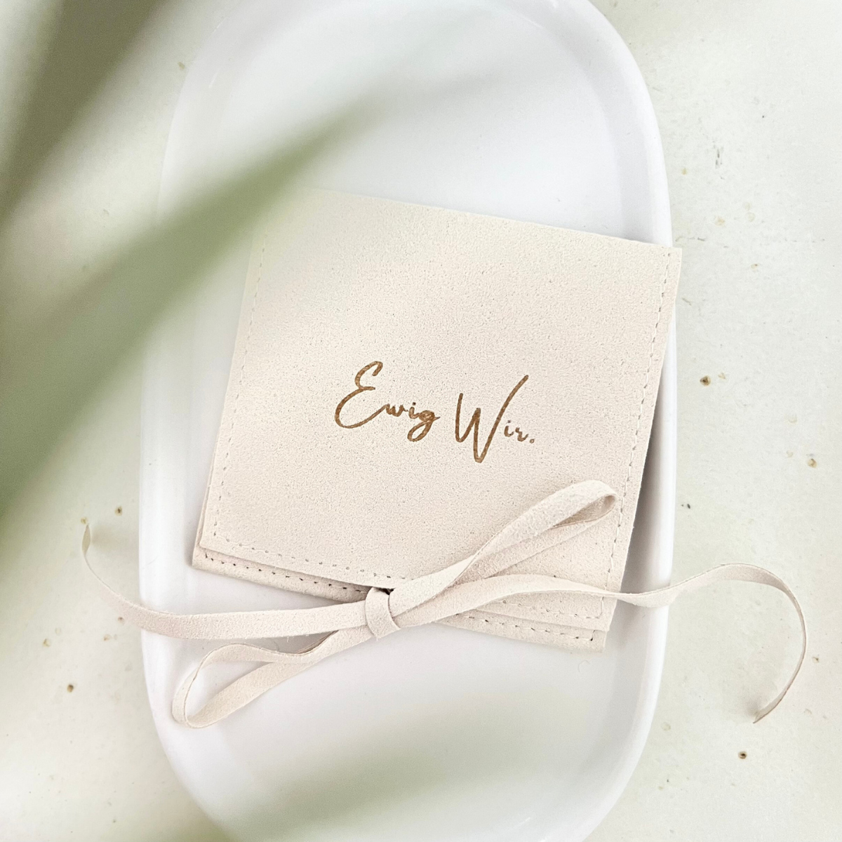 Nahaufnahme eines beigen Schmuckbeutels mit der goldenen Aufschrift „Ewig Wir.“ und Schleife, liegend auf weißer Keramikschale – romantische Geschenkverpackung für Partnerschmuck oder Jahrestagsgeschenke