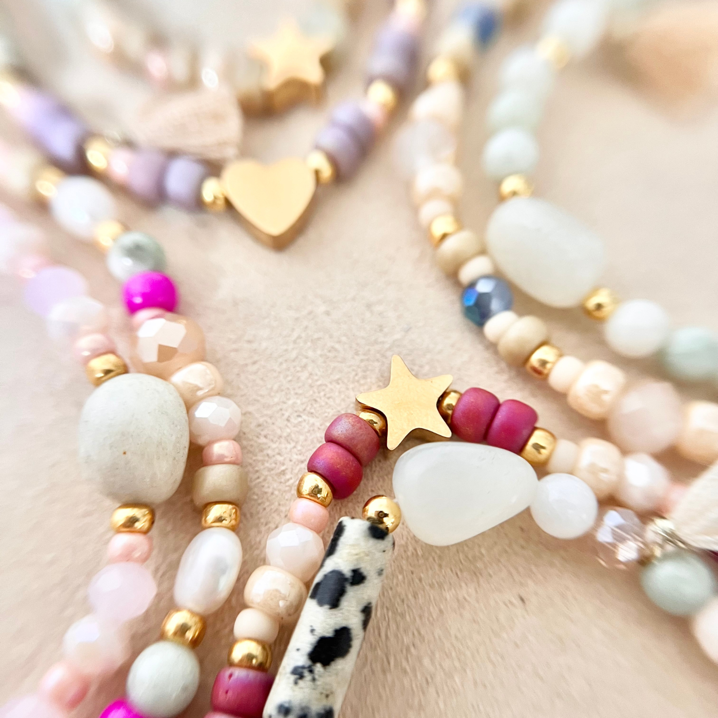 Nahaufnahme mehrerer selbst gestalteter Perlenarmbänder mit unterschiedlich großen Naturstein- und Glasperlen in Creme-, Rosé-, Beere- und Minttönen. Zu sehen sind goldfarbene Edelstahl-Zwischenelemente in Stern- und Herzform, ein schwarz-weiß gemusterter Naturstein sowie matt schimmernde Perlen, arrangiert auf hellem Stoff.