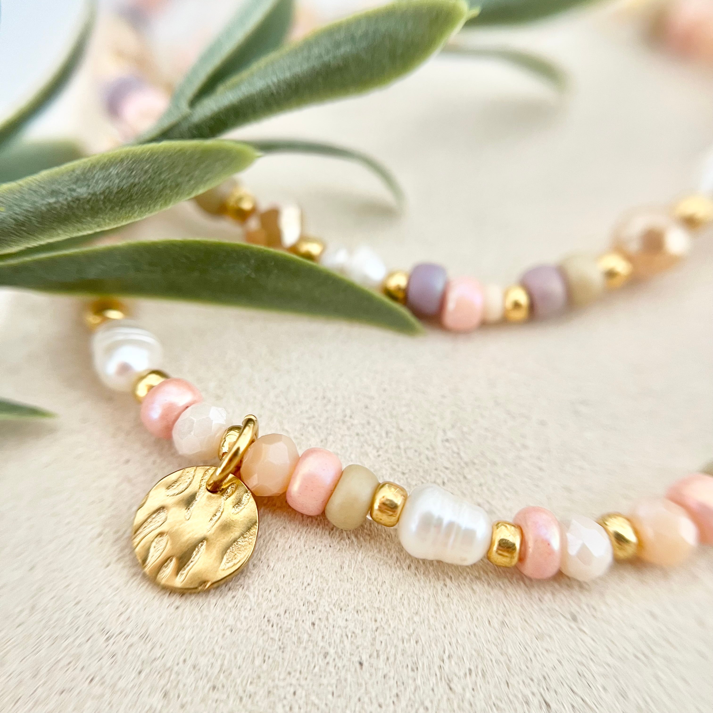 Detailaufnahme eines gehämmerten goldenen Anhängers zwischen zarten Süßwasser- und Glasperlen, umgeben von grünen Blättern.