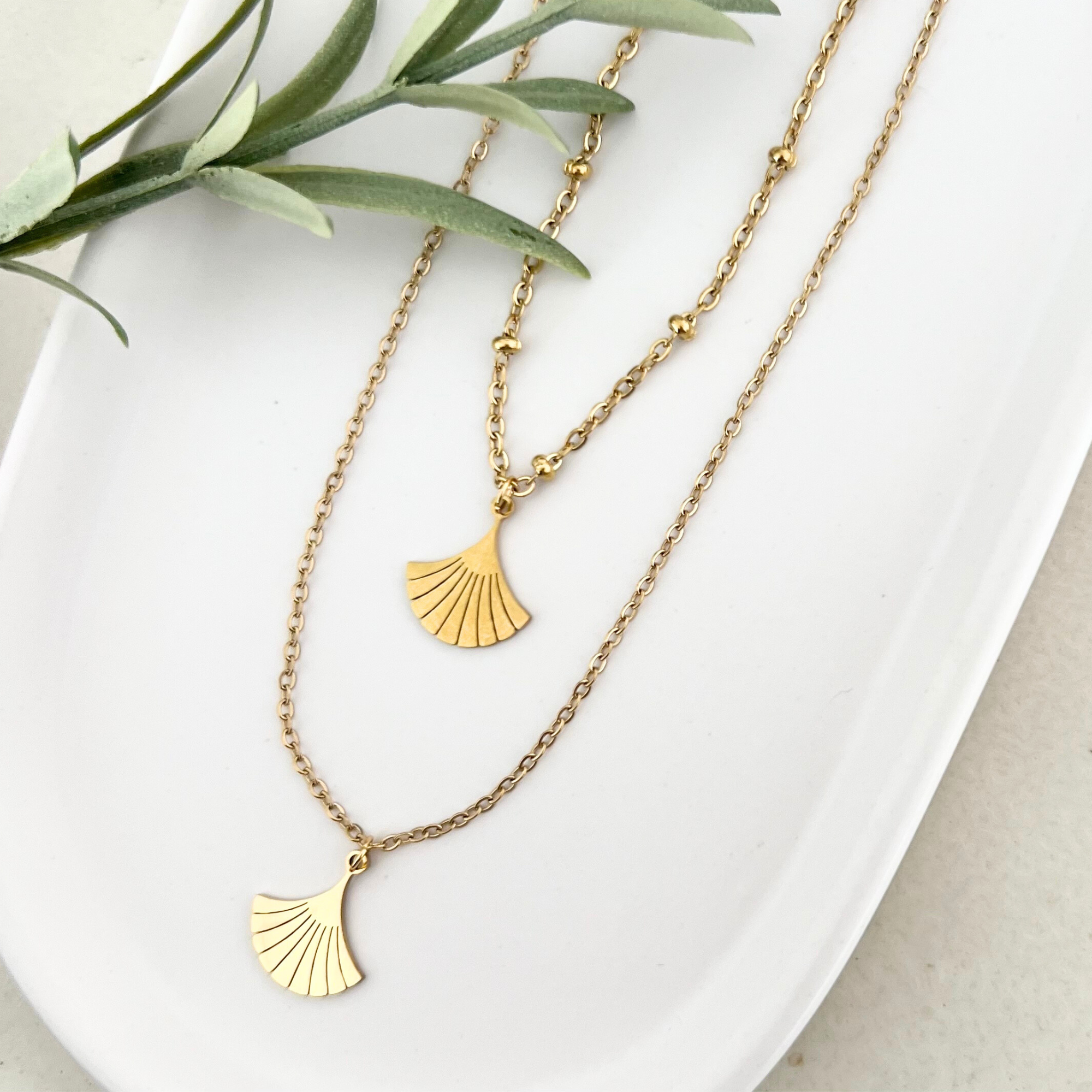 Zwei goldene Edelstahlketten DEIA nebeneinander auf einem weißen Tablet, beide mit filigranen Ginkgo-Blatt Anhängern.