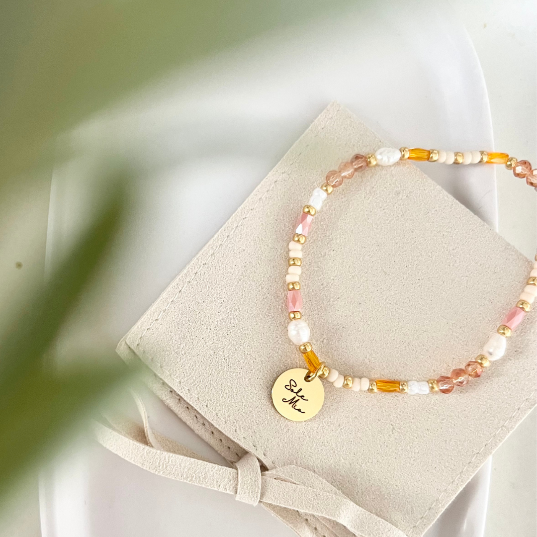 Sommerliches Perlenarmband mit goldenem „Sole Mio“-Anhänger auf hellem Stoffbeutel. Detailaufnahme in Orange-, Weiß- und Goldtönen mit rundem gravierten Edelstahl-Charm.