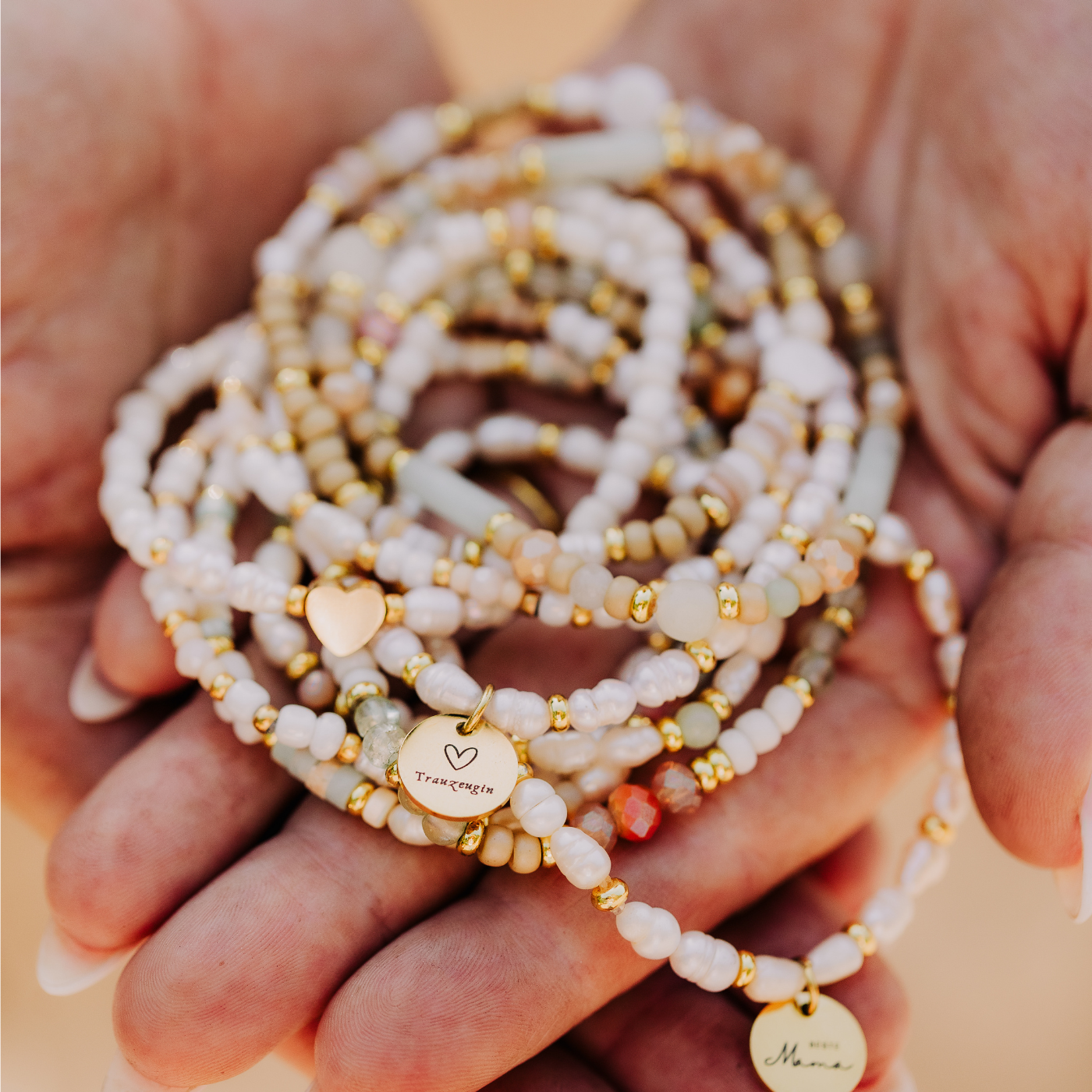 Hand hält mehrere weiße Perlenarmbänder mit goldfarbenen Gravur Anhängern, teilweise mit Herzsymbol und Schriftzügen.