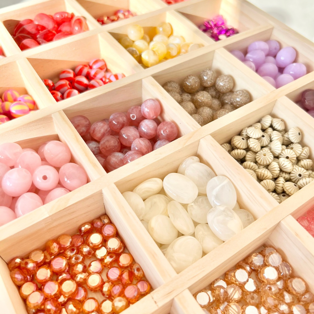 Draufsicht auf eine Holzbox mit mehreren Fächern, gefüllt mit verschiedenen Perlenarten für den Handyketten Workshop, darunter Acrylperlen, Glasperlen und strukturierte Perlen in Rosa-, Rot-, Orange-, Beige- und Lilatönen.