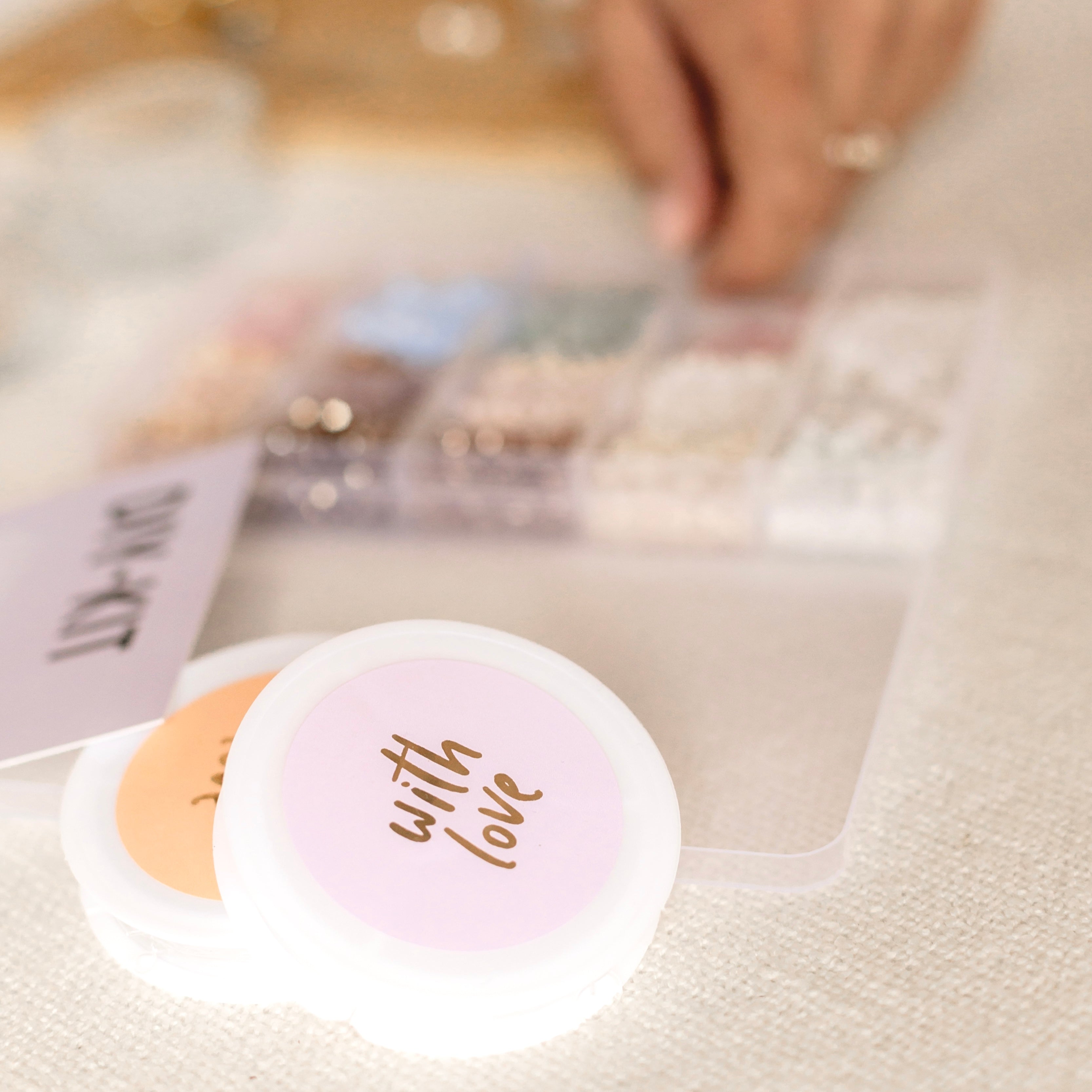 DIY Kit unscharf im Hintergrund, „With Love“-Aufschrift im Vordergrund – kreative Armbänder für unvergessliche Momente.