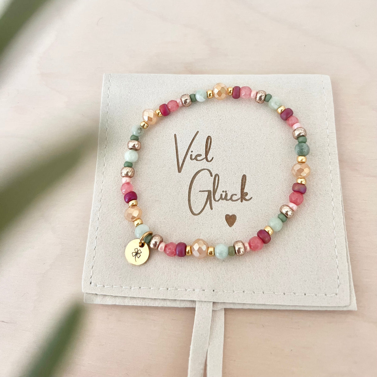 Handgefertigtes Armband FELICIA mit rosa, mintgrünen und goldfarbenen Naturstein- und Glasperlen sowie goldenem Edelstahl-Anhänger mit Kleeblatt-Gravur auf Schmuckbeutel mit „Viel Glück“-Botschaft.