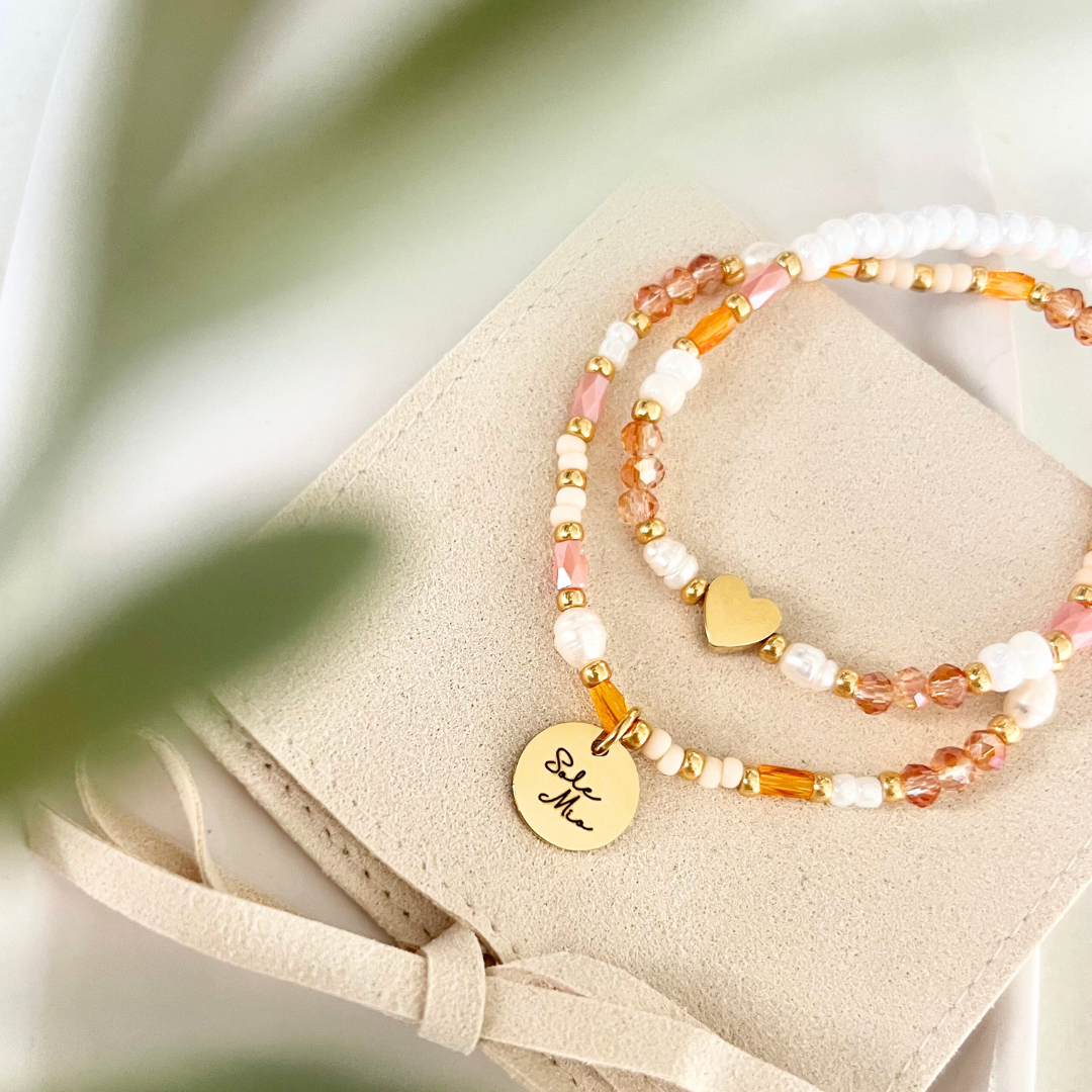 Zwei filigrane Perlenarmbänder in Orange, Weiß und Gold liegen auf einem beigefarbenen Velours-Etui. Mit Süßwasserperlen und Edelstahl Herz Perle. Erinnert an ein Aperol Spritz Armband.