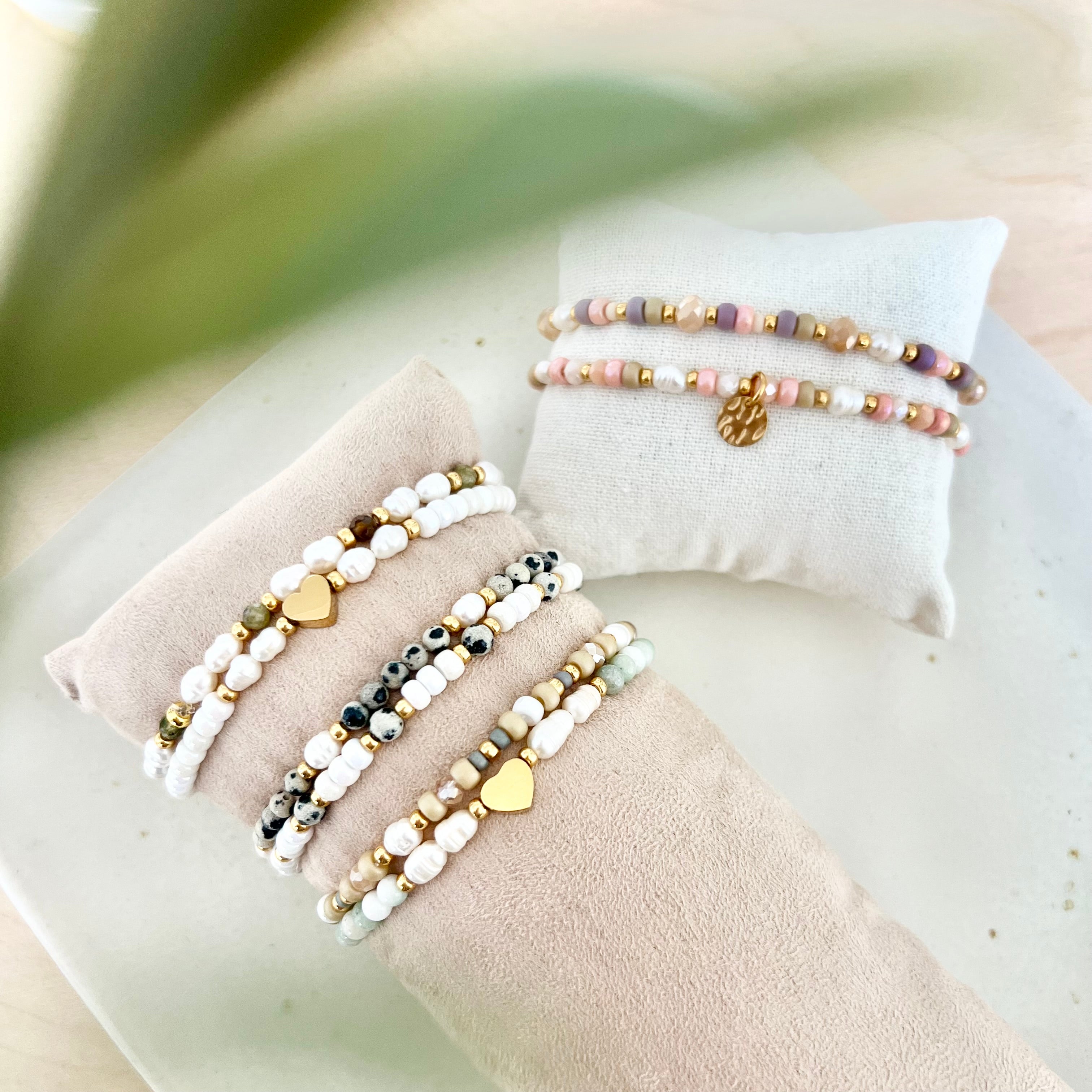 Mehrere handgefertigte Perlenarmbänder in Weiß-, Grau- und Naturtönen mit goldenen Details auf Schmuckkissen arrangiert, sanftes Licht und natürlicher Hintergrund.