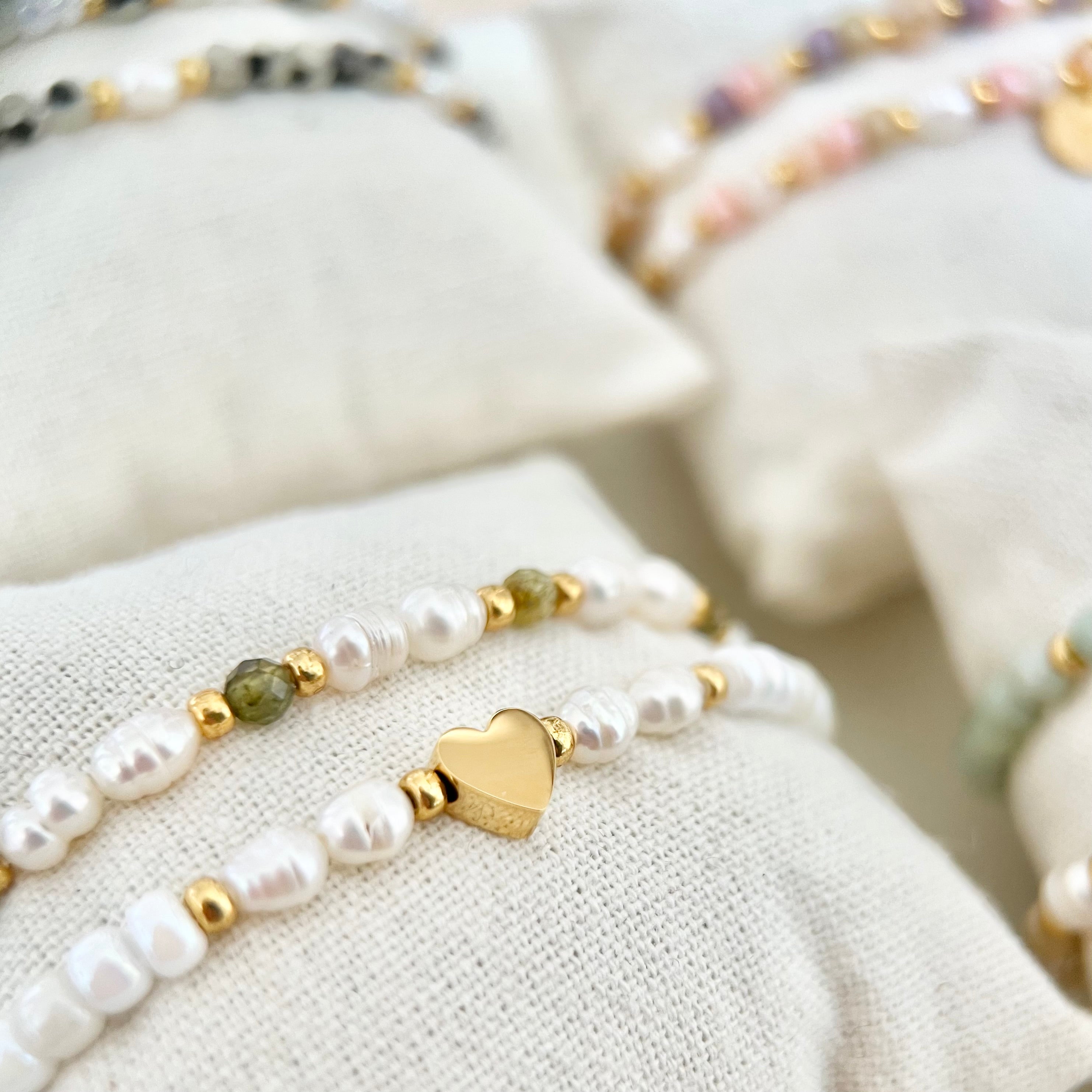 Armbänder in Weiß- und Grüntönen auf Schmuckkissen präsentiert, im Hintergrund zarte Pastellfarben und weiches Licht.