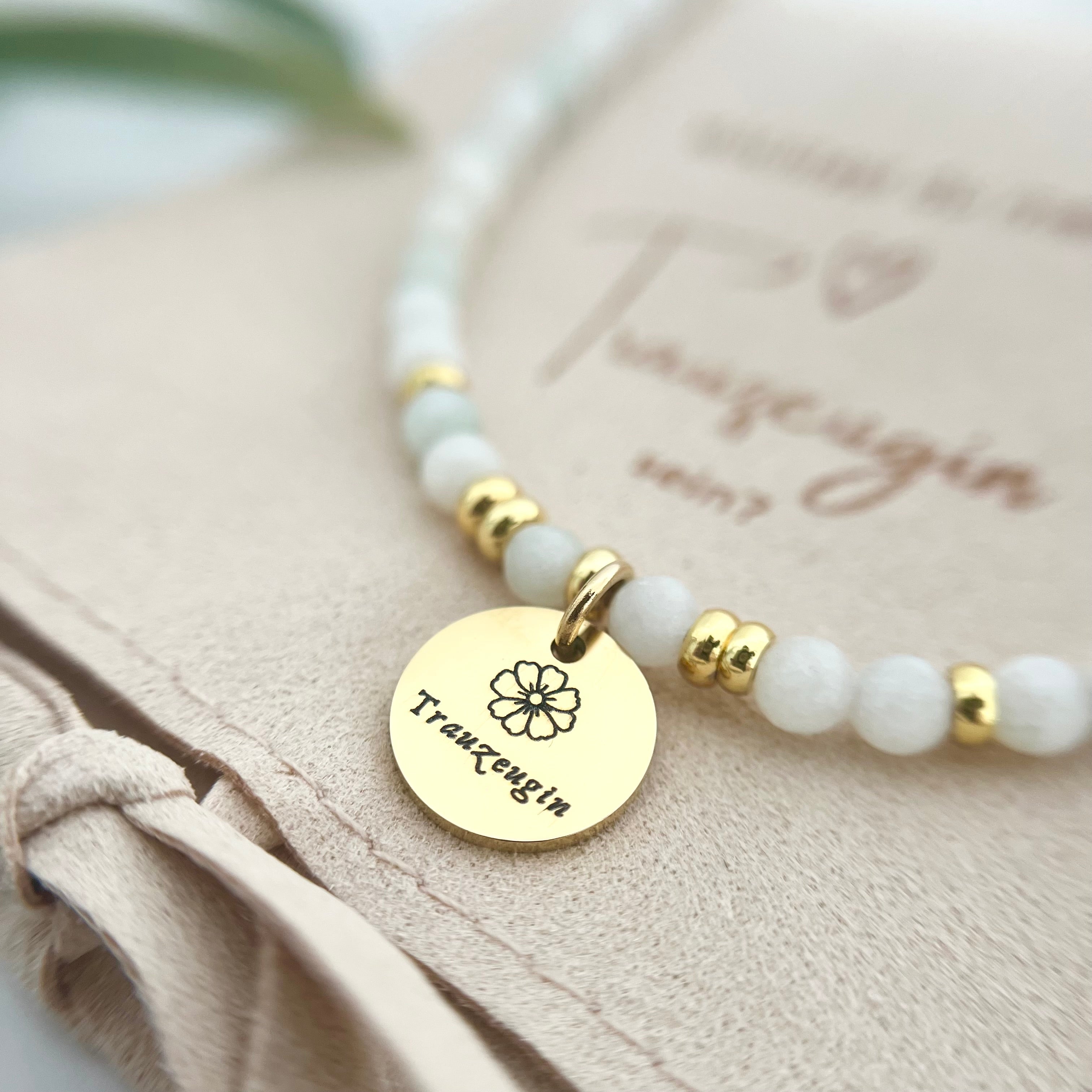 zartes und helles Perlenarmband aus Natursteinperlen mit vergoldetem Edelstahl Anhänger und der Gravur "Trauzeugin". Auf dem Anhänger ist zusätzlich eine kleine Blumen Blüte graviert.