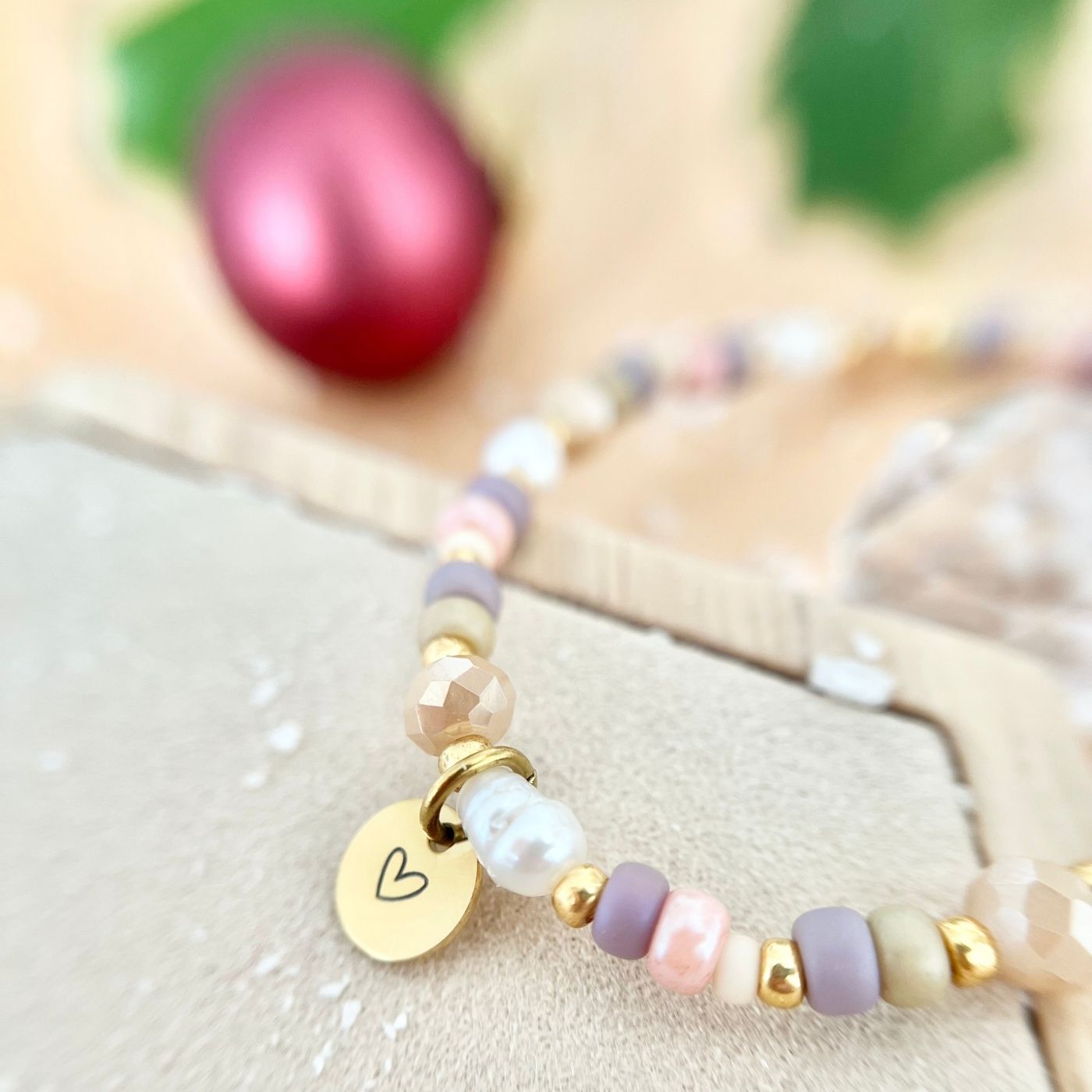 Detailaufnahme eines Armbands in Pastellfarben mit Süßwasserperlen und goldenen Akzenten. Im Fokus ein runder Herzanhänger, unscharfe Weihnachtskugel im Hintergrund.