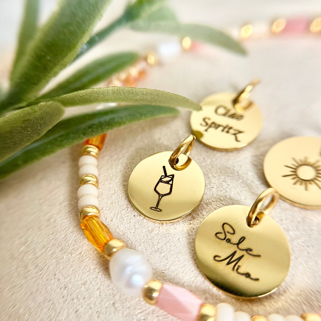 Auswahl an goldenen Gravuranhängern mit Motiven wie Weinglas, Sonne und Schriftzug „Ciao Spritz“. Elegante Edelstahl-Charmanhänger als Ergänzung zum sommerlichen Perlenarmband, inspiriert von Sonne und Genussmomenten.