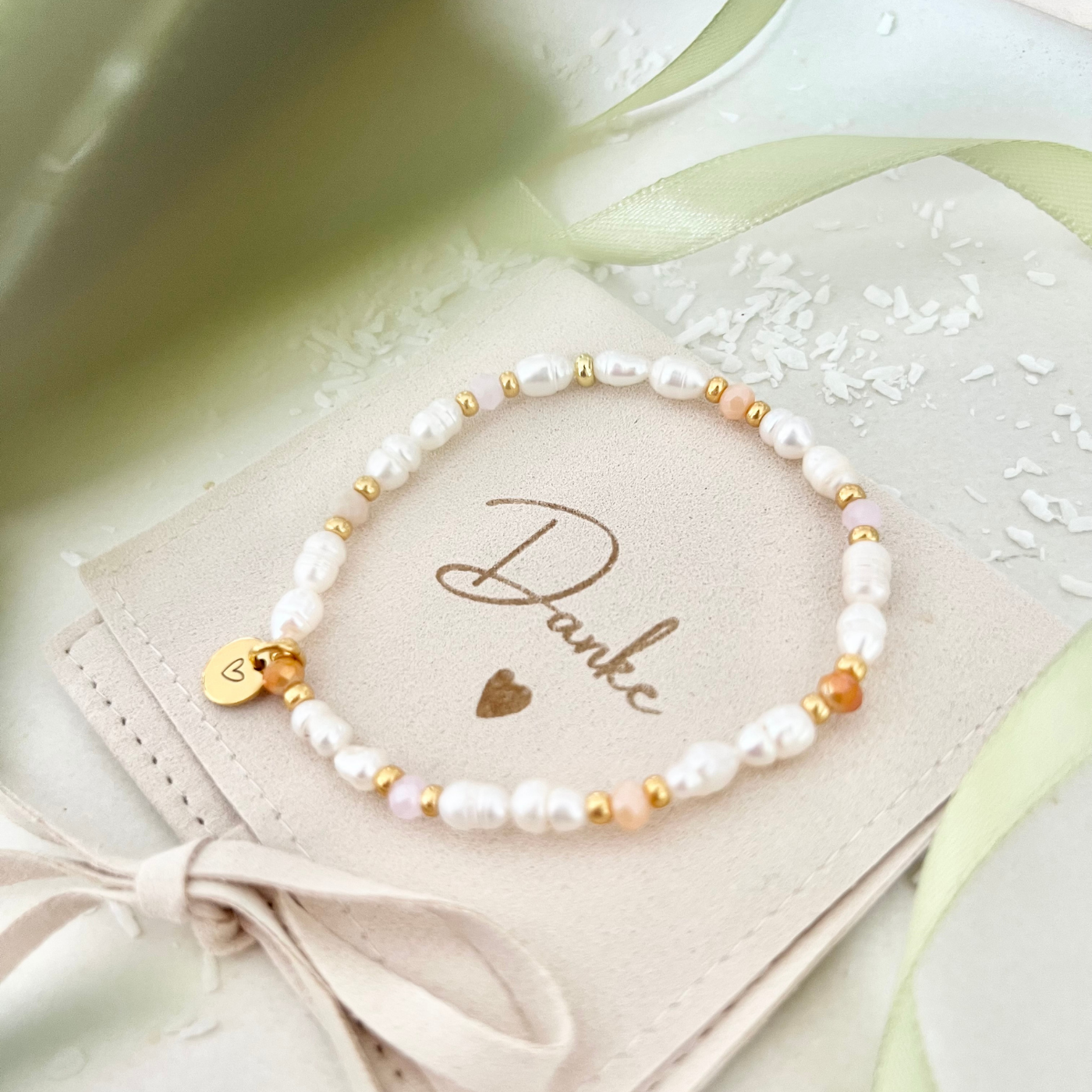 Handgemachtes Perlenarmband aus weißen Süßwasserperlen mit goldfarbenen Zwischenelementen und Herz-Anhänger aus Edelstahl, liebevoll präsentiert auf einem Schmuckbeutel mit der Aufschrift „Danke“ – perfektes Geschenk für besondere Menschen.