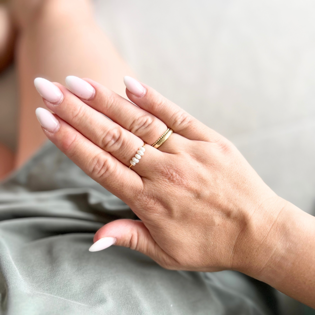 Hand mit filigranem goldenen Ring, verziert mit Süßwasserperlen, kombiniert mit einem weiteren goldenen Ring auf elegant manikürten Fingern, vor einem neutralen Hintergrund – stilvoller Schmuck für feminine Looks, auch als außergewöhnlicher Verlobungsring.