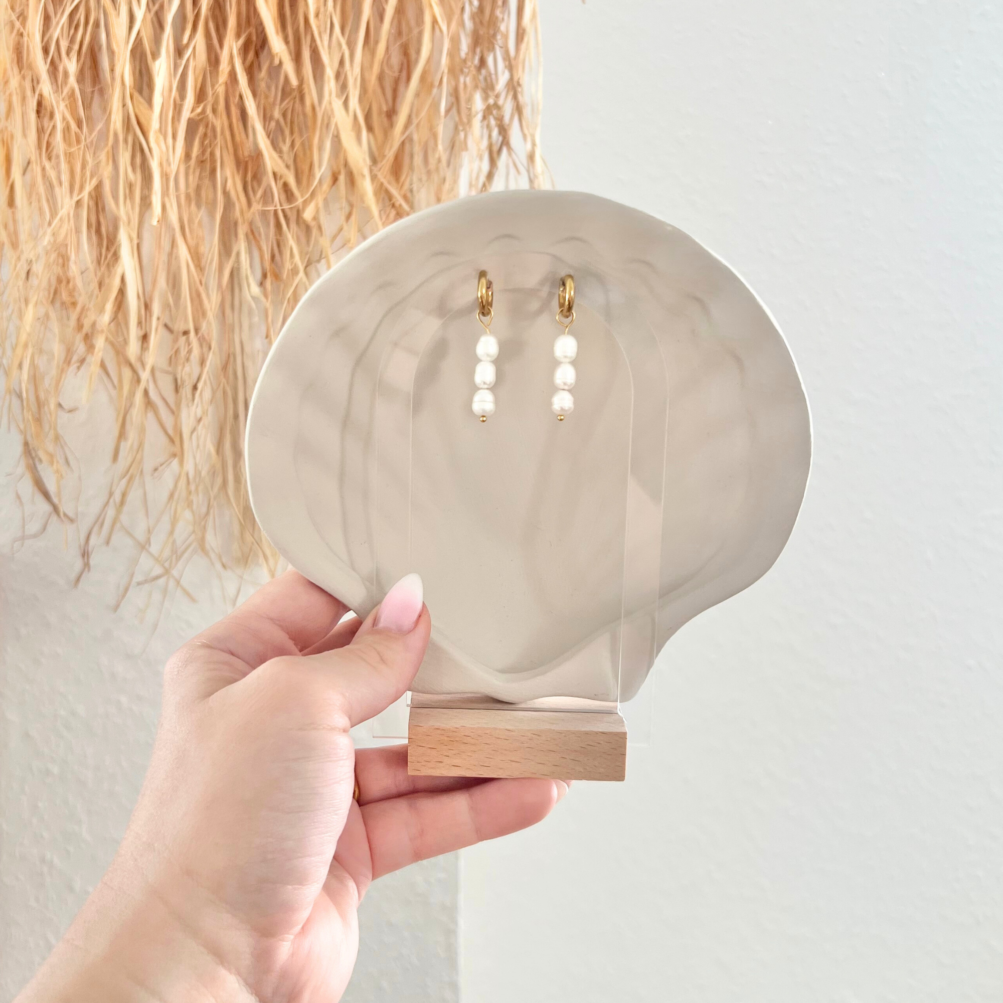 Paar goldene Edelstahl-Ohrringe PERLA mit weißen Süßwasserperlen, auf einem Aufsteller präsentiert, um das elegante Design und die schlichte Raffinesse zu betonen.