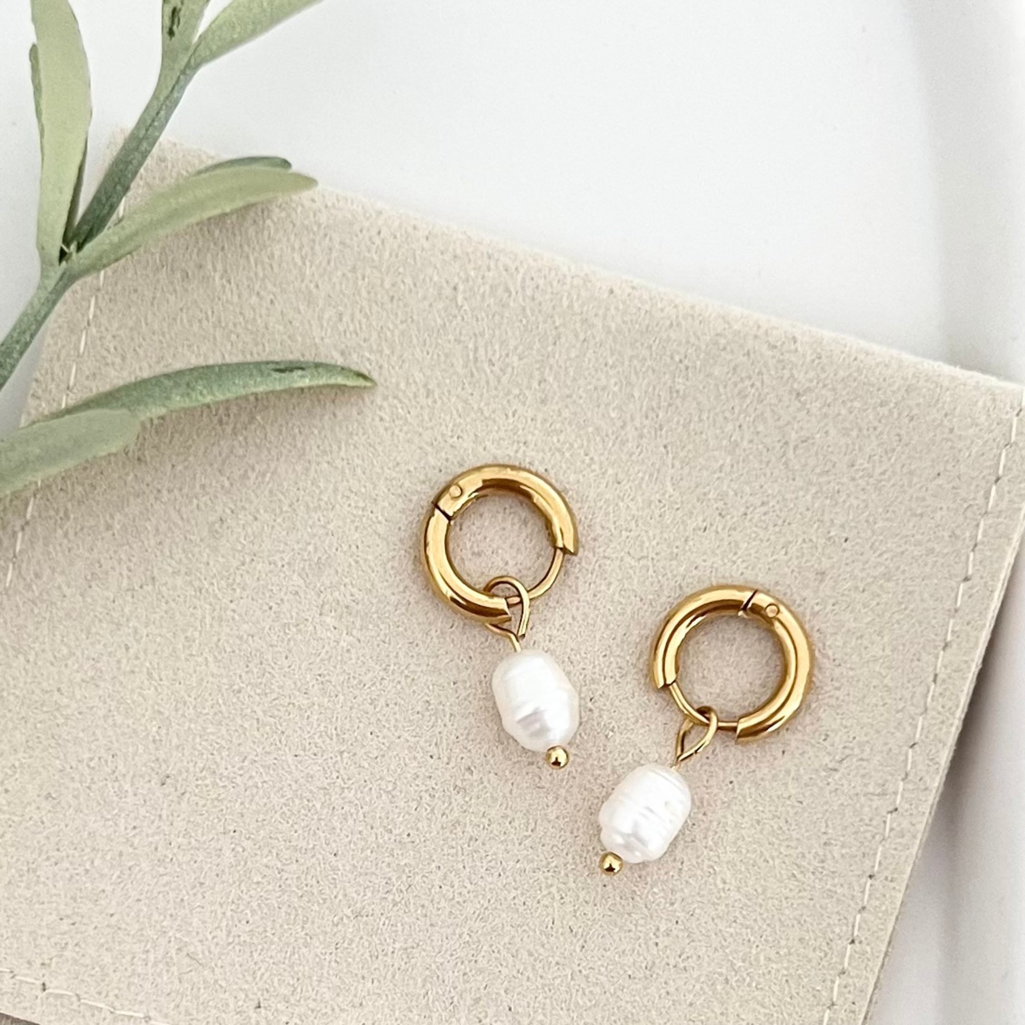Paar goldene Edelstahl-Ohrringe ISLA mit schimmernder Süßwasserzuchtperle, auf einer Tasche drapiert, um das elegante Design und den dezenten Look zu präsentieren.