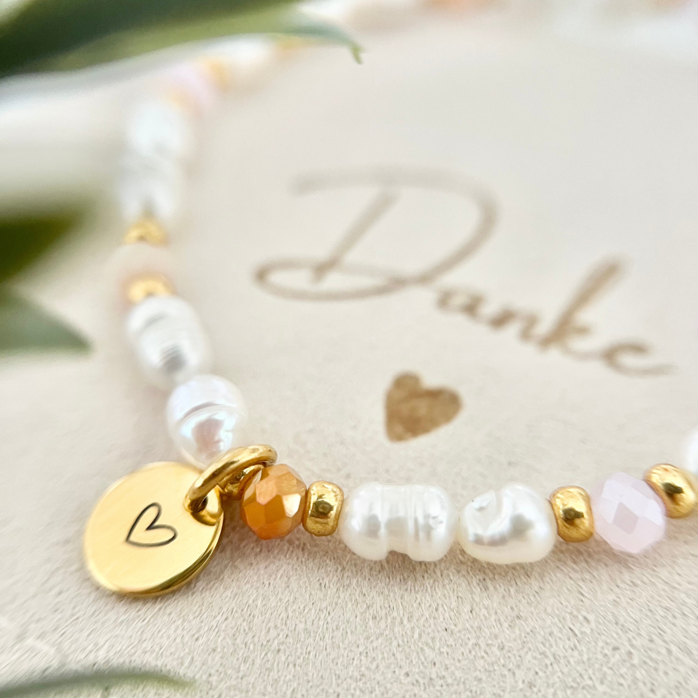 Detailaufnahme eines handgemachten Armbands mit weißen Süßwasserperlen, apricot farbenen Glasperlen, goldfarbenen Zwischenelementen und rundem Edelstahl-Anhänger mit eingraviertem Herz, dekorativ auf Schmuckbeutel mit der Aufschrift „Danke“.