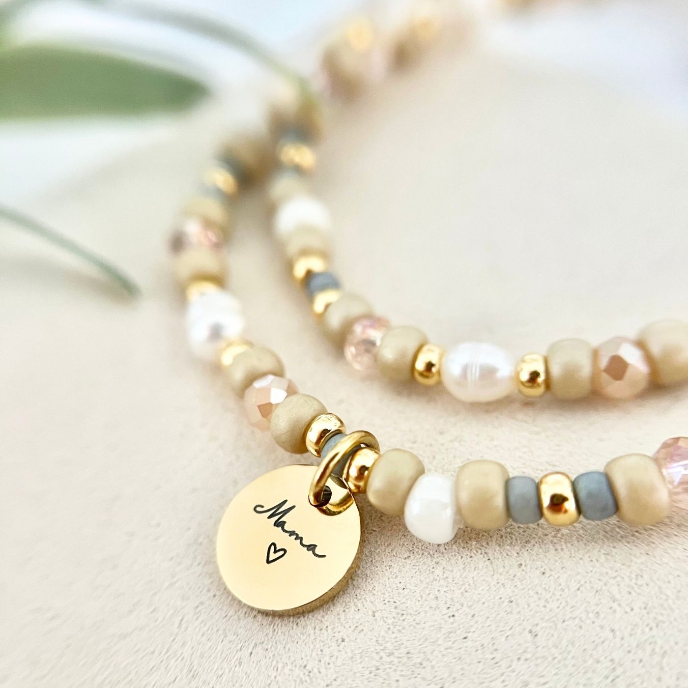 Detailaufnahme eines Armbands mit beigen, weißen und roséfarbenen Perlen, Süßwasserperlen und goldenen Elementen. Gravur-Anhänger „Mama“ mit Herz im Vordergrund.