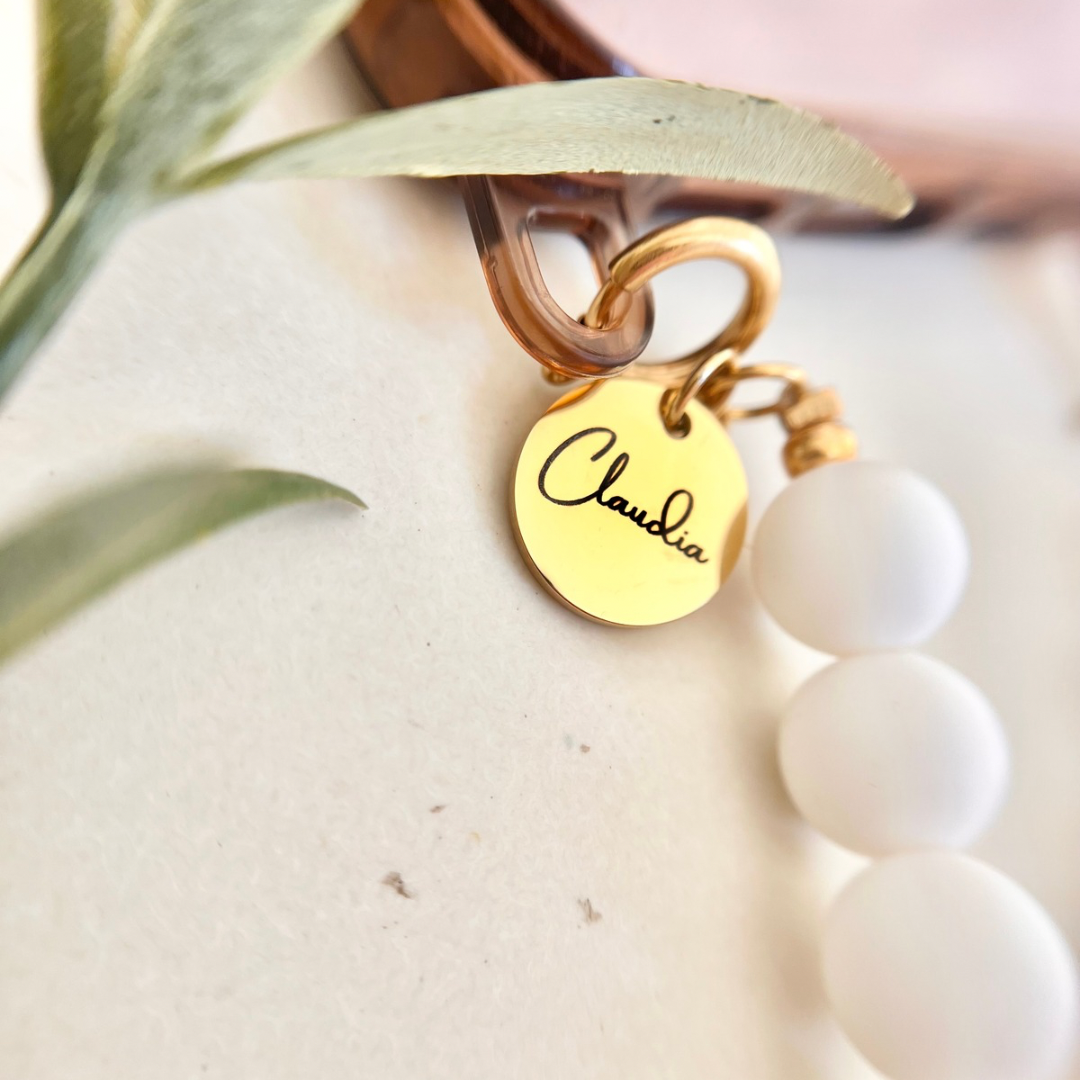 Nahaufnahme einer im Workshop gefertigten Perlen-Handykette mit weißen runden Perlen und einem goldfarbenen Edelstahl-Anhänger mit eingraviertem Namen „Claudia“, befestigt an einem Smartphone mit transparenter Hülle und goldenem Karabinerring, neben grünen Pflanzenblättern.