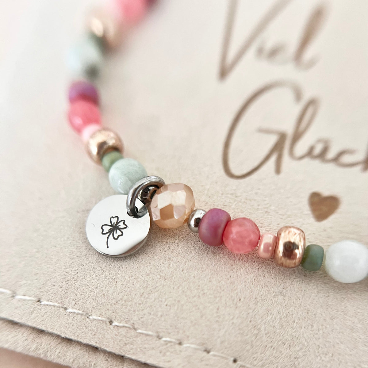 Detailaufnahme des handgefertigten Armbands FELICIA in Rosa, Mint und Roségold mit Naturstein- und Glasperlen sowie silberfarbenem Edelstahl-Anhänger mit graviertem Kleeblatt, auf einem Schmuckbeutel mit „Viel Glück“-Botschaft.