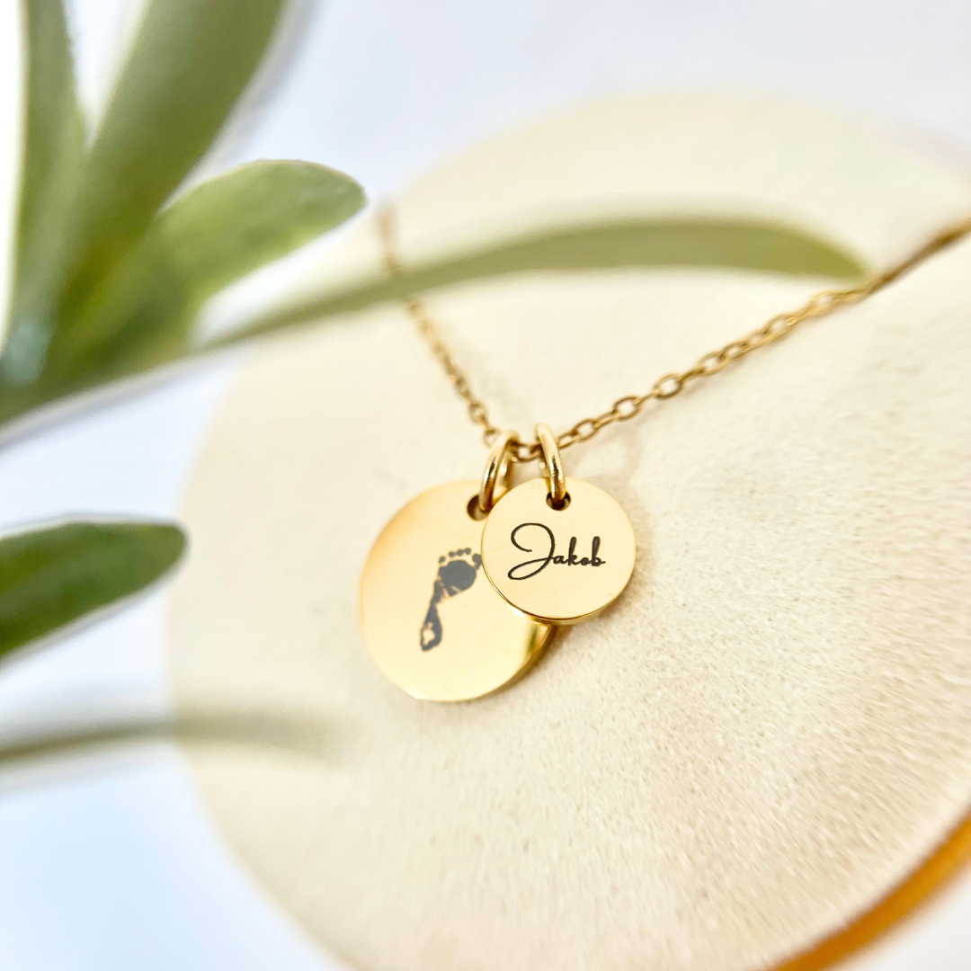 Goldene Gravur Kette mit zwei runden Anhängern – einer mit eingraviertem Babyfuß, der andere mit dem Namen „Jakob“. Personalisiertes Schmuckstück aus Edelstahl.