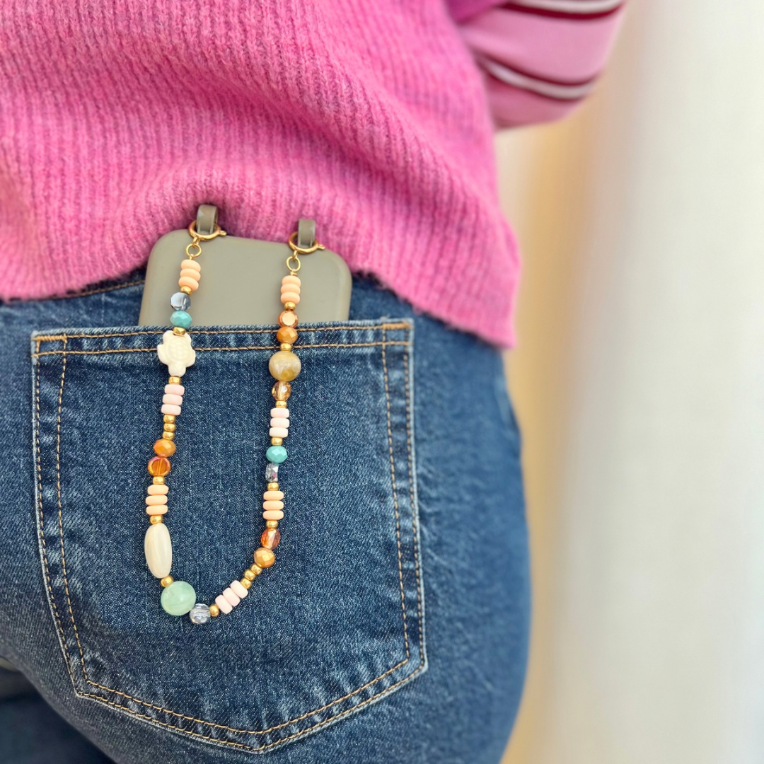 Person trägt eine im Handyketten Workshop gefertigte kurze Perlen-Handykette mit bunten Glas-, Acryl- und Natursteinperlen in Rosa-, Türkis-, Beige- und Orangetönen, befestigt an einem Smartphone, das in der Gesäßtasche einer Jeans steckt.